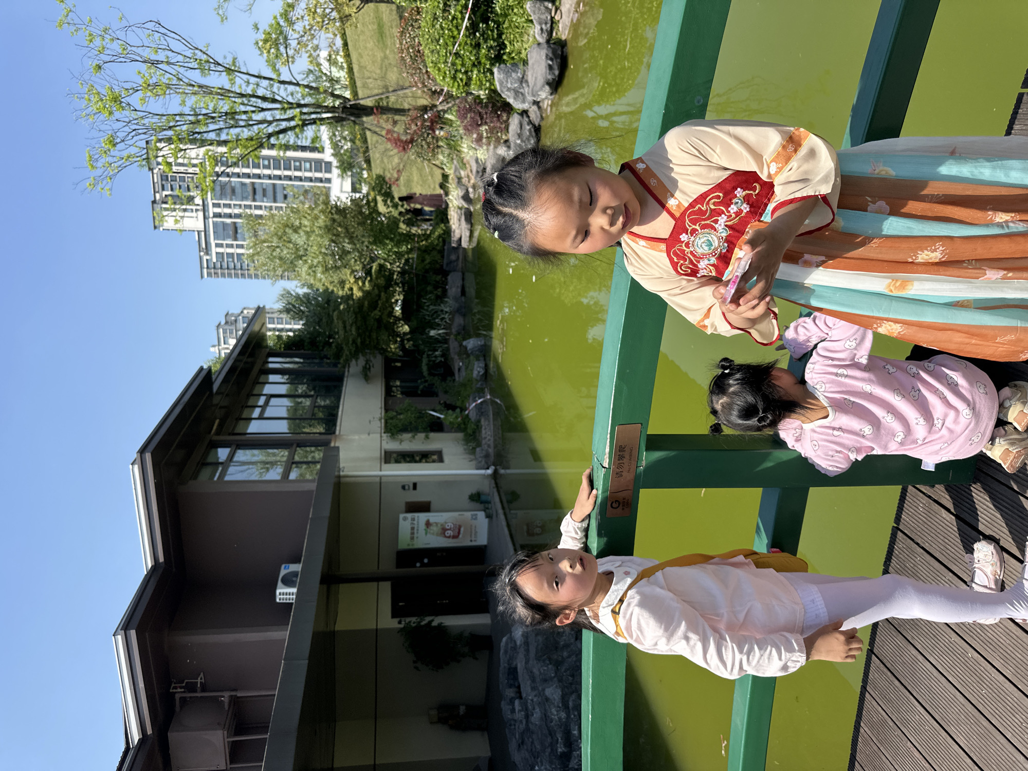 Three young girls are on a green painted wooden bridge overlooking a greenish pond. One girl wears a traditional Chinese dress, while another wears pink pajamas. A third girl wears white leggings and a white top with a yellow strap. The background includes a modern building, trees, and a clear blue sky. The city is identified as Jiangjin, China.
