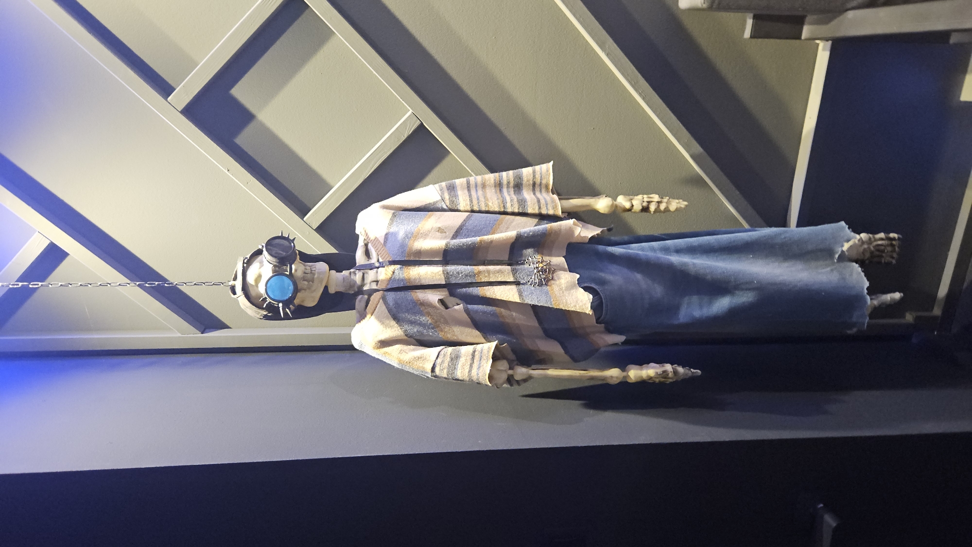 A full skeleton, dressed in a striped poncho, blue denim skirt, and steampunk goggles, hangs from a chain against a wall with a diamond pattern. The skeleton has its mouth open and appears to be mid-sentence, with its eyes closed. A large, ornate necklace dangles from its neck. The background is a dim, industrial-looking room, possibly a bar or club, with a blue light emanating from the top left of the frame.