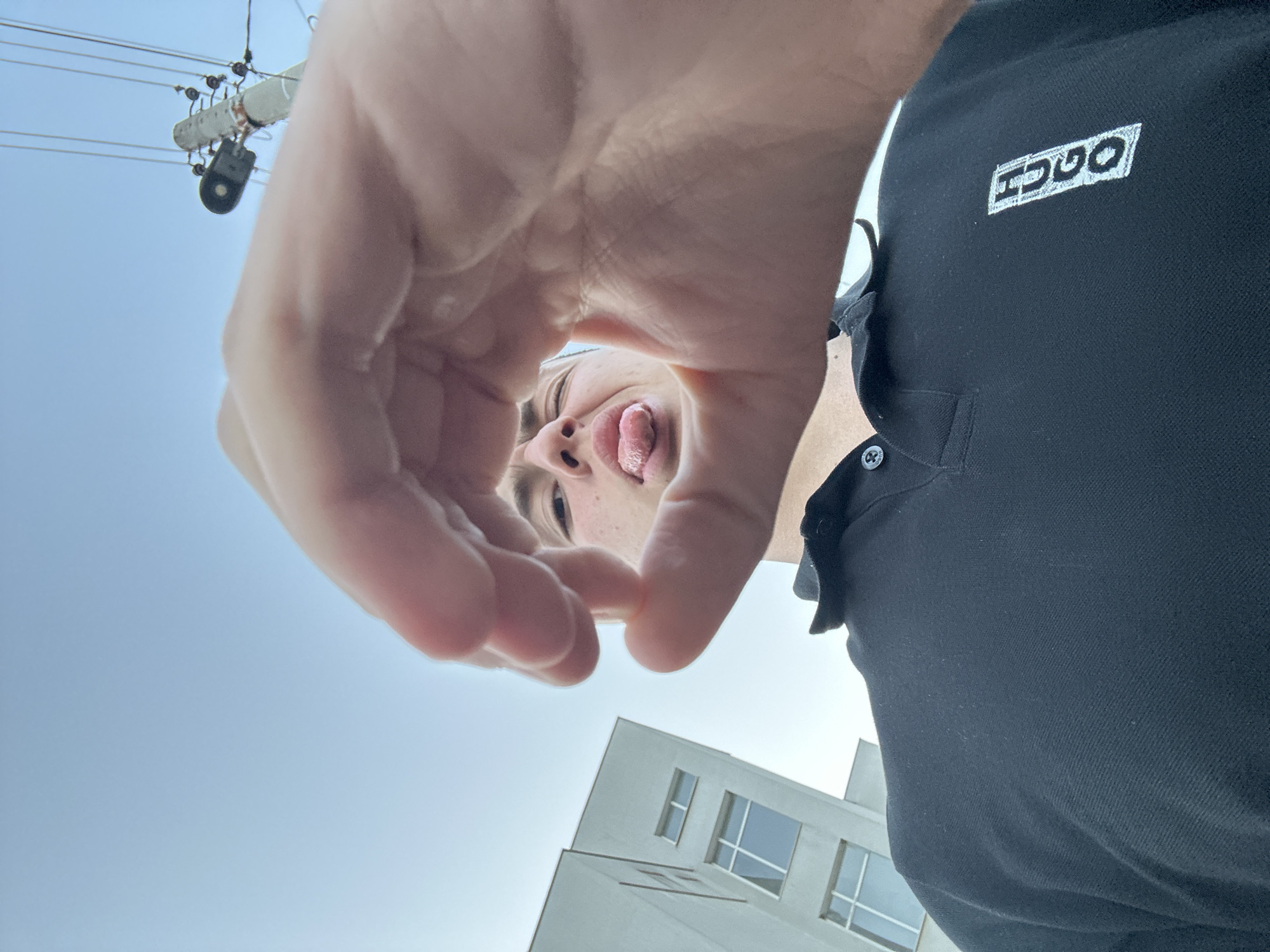 A low-angle photograph depicts a person with their tongue sticking out, framed by their hand in the foreground. They are wearing a black collared shirt with the word "HUGO" on the left breast. Above and to the left of the person are electrical lines connected to a pole. In the background is a modern building with multiple windows and a white exterior. Based on the architectural style and infrastructure, the image was likely taken in an urban environment. It's difficult to pinpoint a specific location in Zlatograd, Bulgaria, from this image alone.
