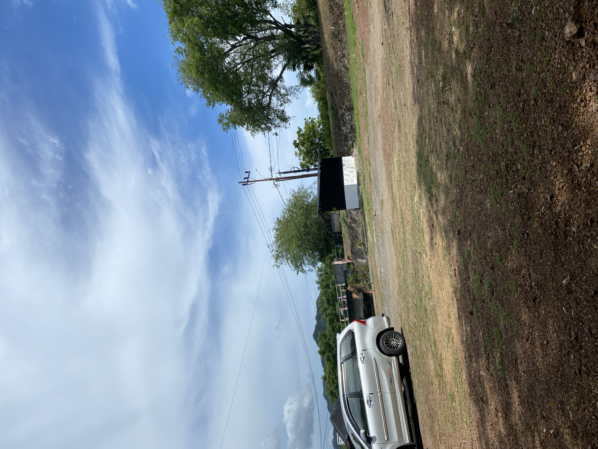 This image depicts a roadside scene in Aneuk Paya, Indonesia, under a partly cloudy sky. In the foreground, a silver minivan is parked on a gravel and grass area. The vehicle shows a "TRD SPORTIVO" decal on its side. Behind the car, a small, dark-roofed structure with white walls is visible, situated on a slightly elevated embankment. A utility pole with electrical wires stands near this structure. Beyond the immediate foreground, a bridge spans a waterway, with trees and hills forming the backdrop. The overall environment appears to be a rural or semi-rural setting.