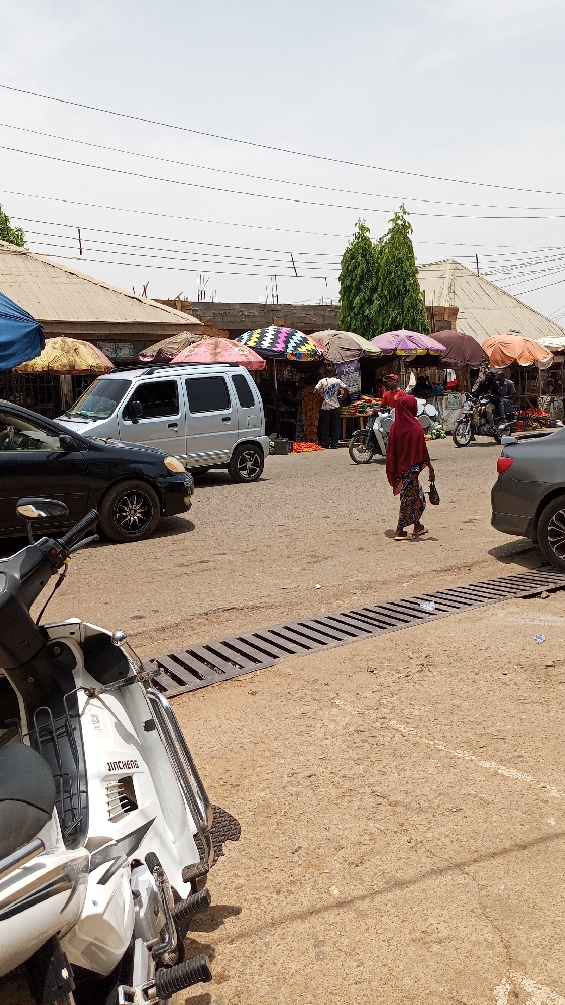 The image captures a vibrant street market scene in Narayi, Nigeria, on a bright, hazy day. The setting is an active commercial area with a dusty, light-brown road, partially covered by a metal drainage grate in the foreground.
In the midground, a person wearing a red headscarf and a patterned skirt walks across the street, carrying a small item. Vehicles are present; a black sedan is partially visible on the left, and a silver compact SUV-like vehicle is either parked or moving slowly. Several market stalls line the side of the road, each sheltered by a colorful umbrella – shades of pink, purple, brown, and a distinctive black-and-white zigzag pattern. Vendors and shoppers are visible around these stalls, with fresh produce, including red tomatoes, displayed. Motorcycles are also part of the street activity, with one parked near the stalls and another partially visible on the far right.
The background reveals a mix of buildings, some with corrugated metal roofs, and signs of ongoing construction with visible rebar. Tall, slender green trees rise above the structures. The bright, clear sky is crisscrossed by numerous utility wires.
Visible text includes "JINCHENG" on the white motorcycle in the foreground. At the market stalls, a white sign displays "POS" (Point of Sale), and faint logos for "OPay" and "Mastercard" are discernible on another banner. "CONGAI" is partially visible on a purple umbrella. The overall scene depicts a bustling local market under a midday sun.