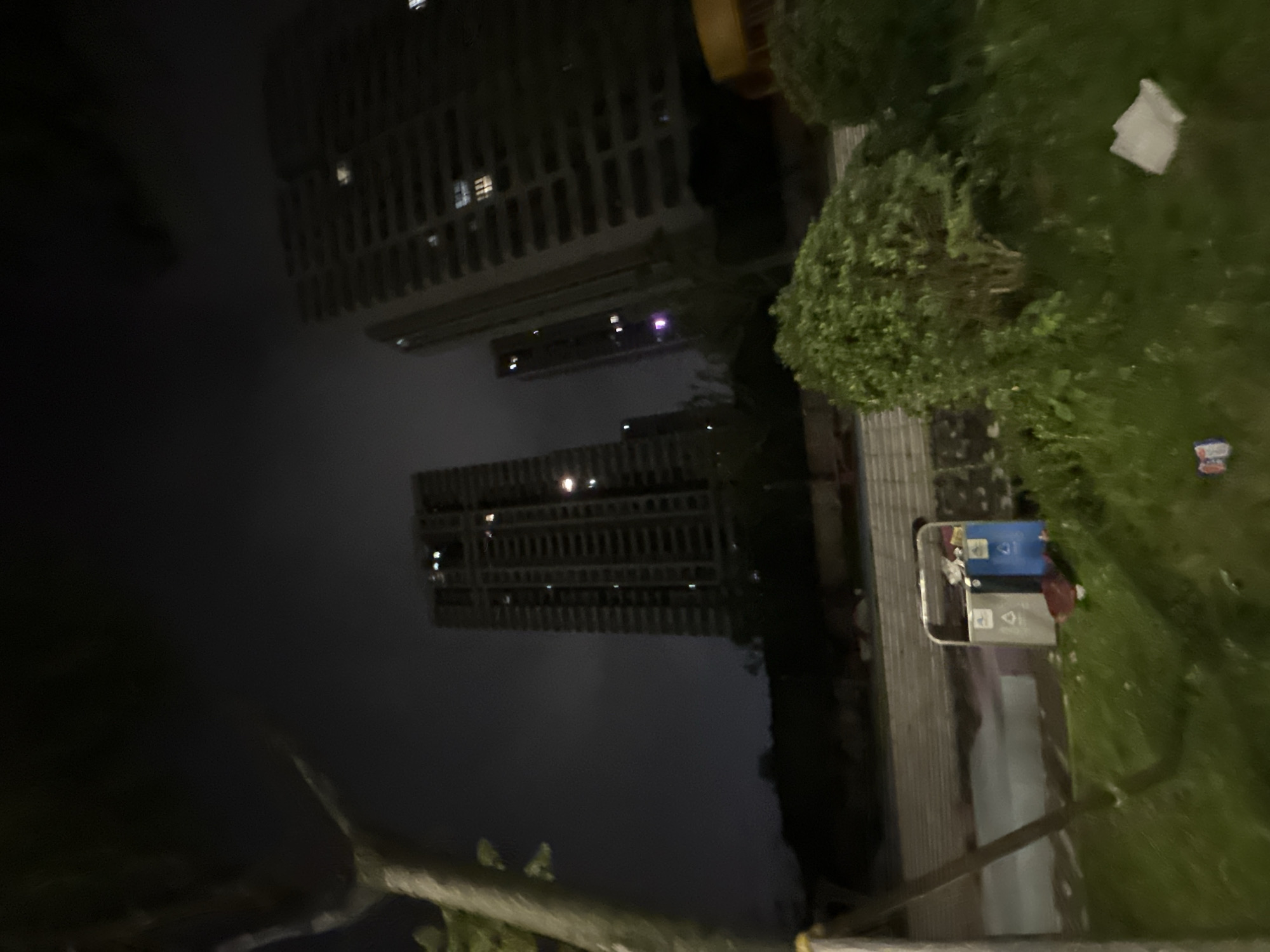 The image depicts an outdoor scene at night, likely in an urban environment. In the background, there are tall, modern buildings with lit windows, suggesting a developed area. The foreground features a grassy area with some greenery and a trash receptacle. The lighting is dim, and the overall image has a somewhat blurry quality, characteristic of low-light photography. The scene appears to be located in the Chongqing High-tech Zone, China, as indicated by the context provided. There are no visible people in the image.
