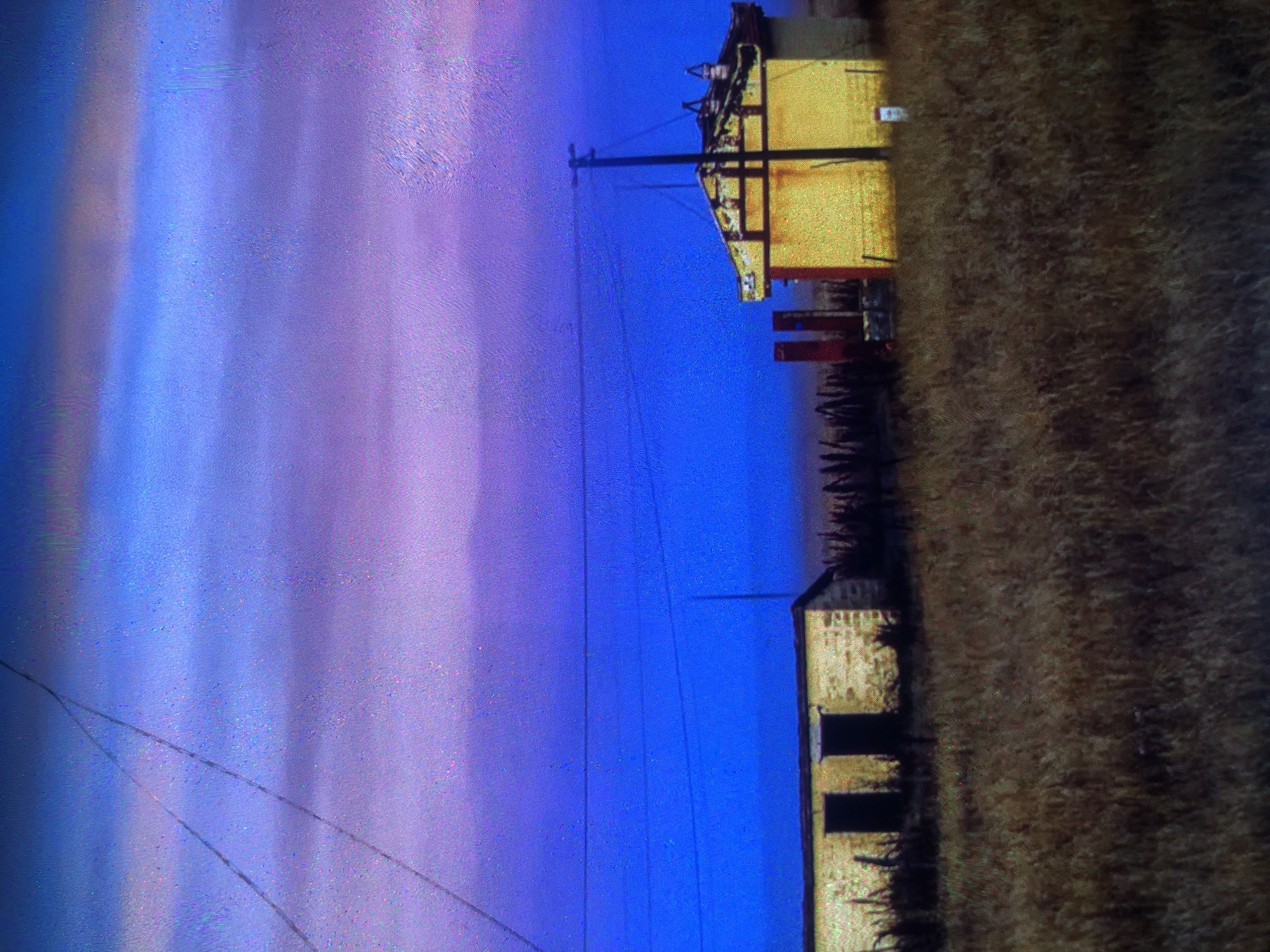 The image depicts a rural landscape at dusk, with a striking sky transitioning from deep blue to shades of purple and pink. In the foreground, dry, golden grass covers the ground. To the left, a small, rectangular building constructed of light-colored stone with two dark doorways is visible. To the right, a taller, yellow building with a red-trimmed roof and a dark pole with wires extending from it stands. Several other structures, including two red pillars, are situated between the two buildings. The overall impression is one of a serene, isolated setting under a vibrant twilight sky. There is no indication that this scene is located in Tashkent, Uzbekistan.
