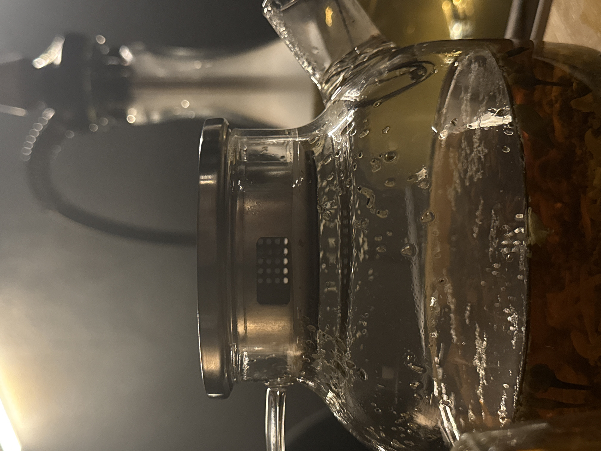 The image is a close-up shot of a glass teapot filled with a dark liquid, likely tea, with some sediment at the bottom. The teapot has a metal lid and a metal handle attached to the side. Condensation is visible on the exterior of the glass. In the background, out of focus, is another glass object, possibly a hookah or a different type of vessel, illuminated by soft, warm lighting. The overall ambiance is dimly lit and atmospheric. The scene is indoors, with no discernible elements that would place it specifically in Barcelona, Spain.