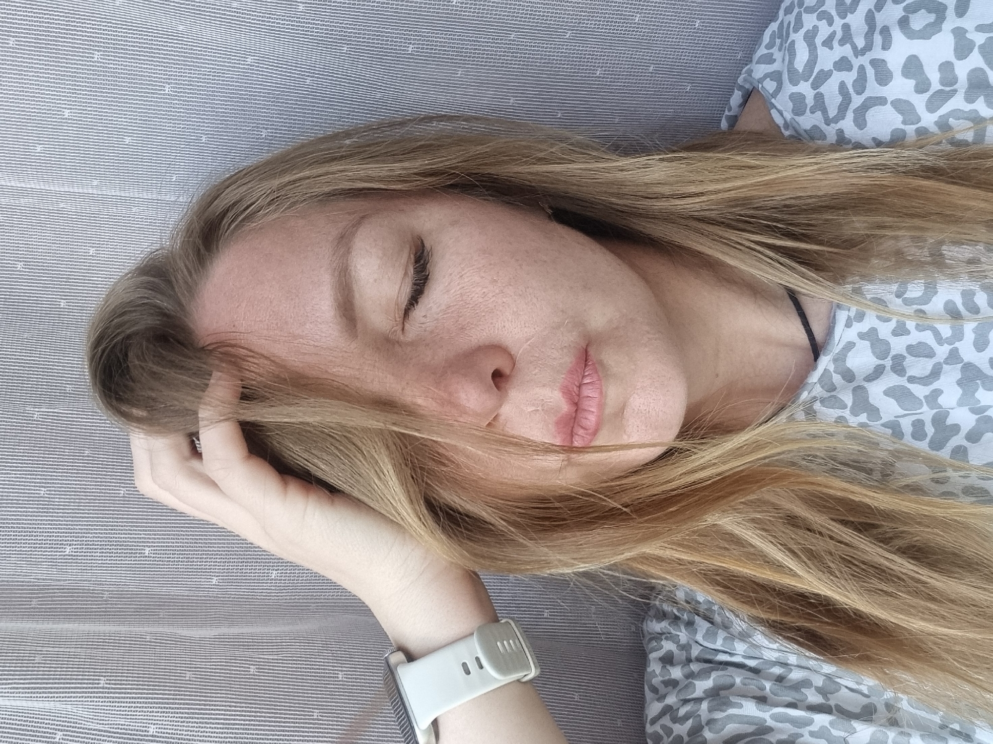 A woman is lying down, eyes closed, with her head resting on her left hand. She is wearing a light-colored shirt with a gray pattern, and a white smartwatch on her left wrist. Her long, blonde hair partially covers her face. She is lying on a gray surface with a textured pattern. The location is not discernible from the image.