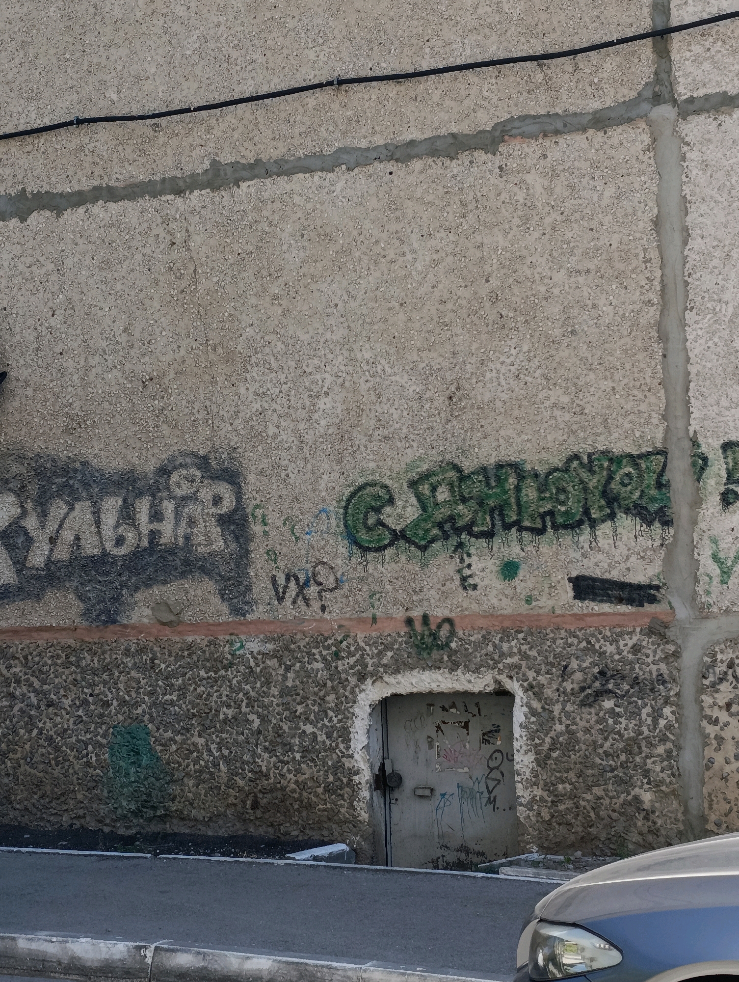 The image shows a section of a concrete building wall with graffiti. The graffiti is in Russian and appears to be spray-painted. One piece of graffiti, in dark gray, reads "КУЛЬТПРОСВЕТ". Another piece, in green with dripping paint, reads "СВЕТЛОКОР". Below the graffiti is a recessed doorway with a metal door covered in various markings and graffiti. The wall has a rough, textured surface composed of small pebbles embedded in concrete. A thin horizontal strip of reddish-brown paint or material runs across the wall below the main graffiti. The building is located in the city of Kostanay, Kazakhstan. The lower part of the image shows a section of a paved road and a blue car.