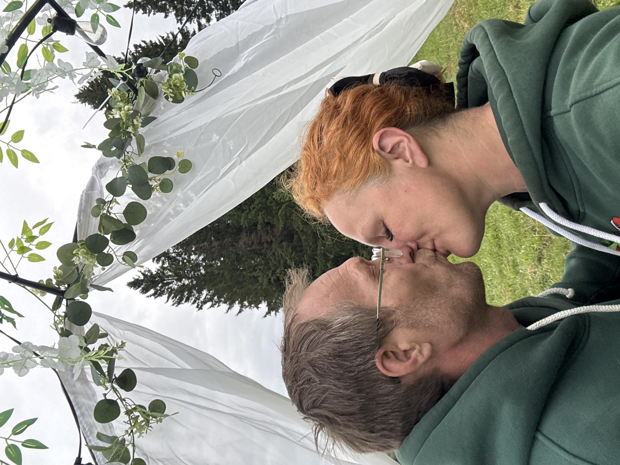 A side-profile shot captures two adults, a male and a female, kissing outdoors. The male, positioned on the left, has brown-grey, wind-swept hair, a faint stubble, and wears thin-framed glasses with gold-colored frames. The female, on the right, has red-orange hair pulled back, and her eyelids are closed during the intimate embrace. Both individuals are wearing matching dark green hooded sweatshirts with white drawstrings. They are situated in a grassy area, beneath a white fabric arch draped over dark poles. The arch is decorated with artificial green foliage, including eucalyptus-like leaves and smaller green clusters, interspersed with white wisteria-style flowers. A single vintage-style clear light bulb hangs from the arch on the upper right. In the background, beyond the arch, several tall, dark evergreen trees stand against an overcast, light grey sky. The scene is located in the city of Bol'shaya Kivara, Russia.