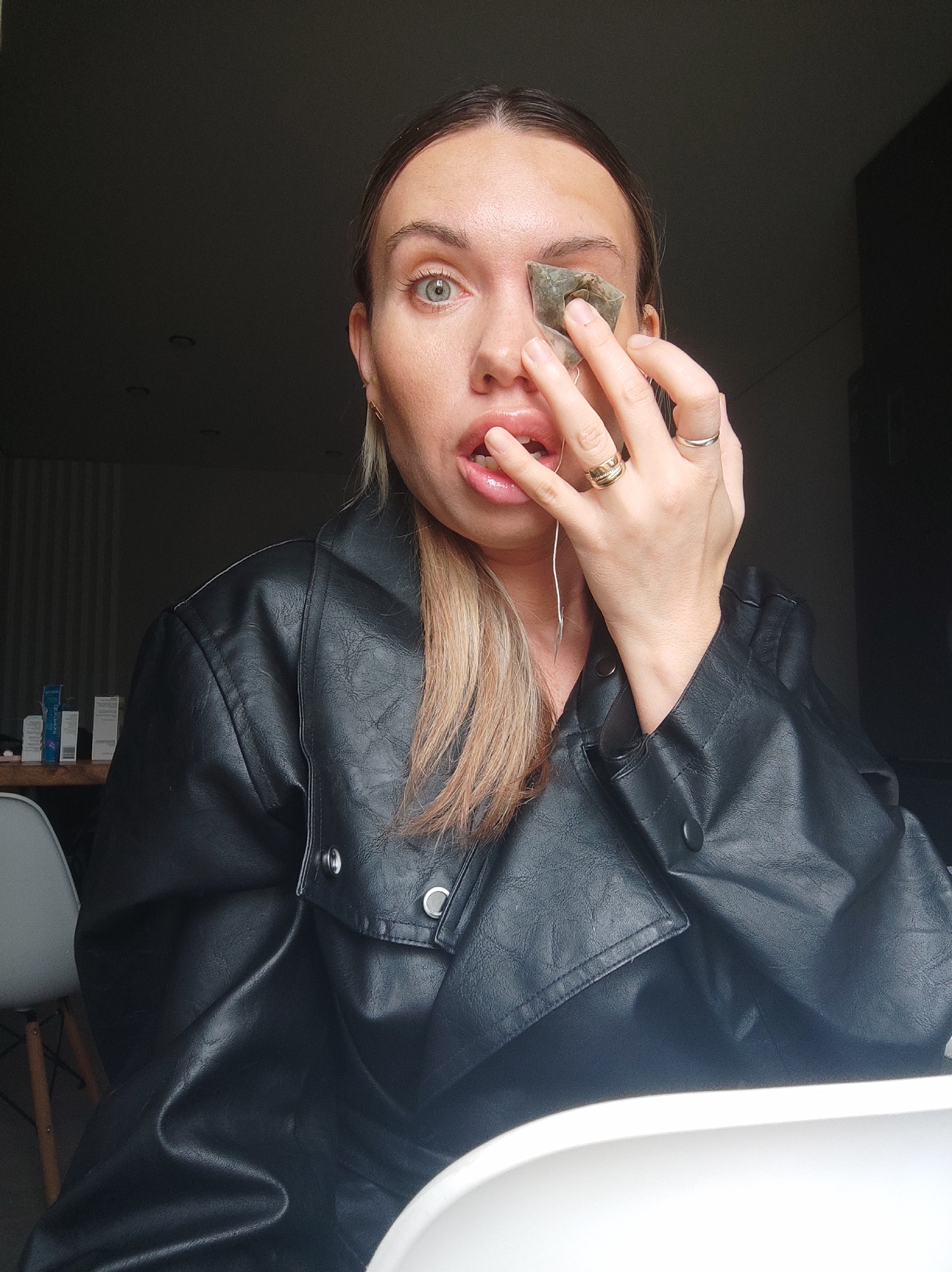 A woman is indoors, holding a tea bag to her left eye. She is wearing a black leather jacket and several rings. The background is a dimly lit interior with a glimpse of what appears to be a desk with cosmetic items and a chair. The location is unknown and not evidently recognizable as the city of Votkinsk, Russia. The action is the woman applying a tea bag to her eye.