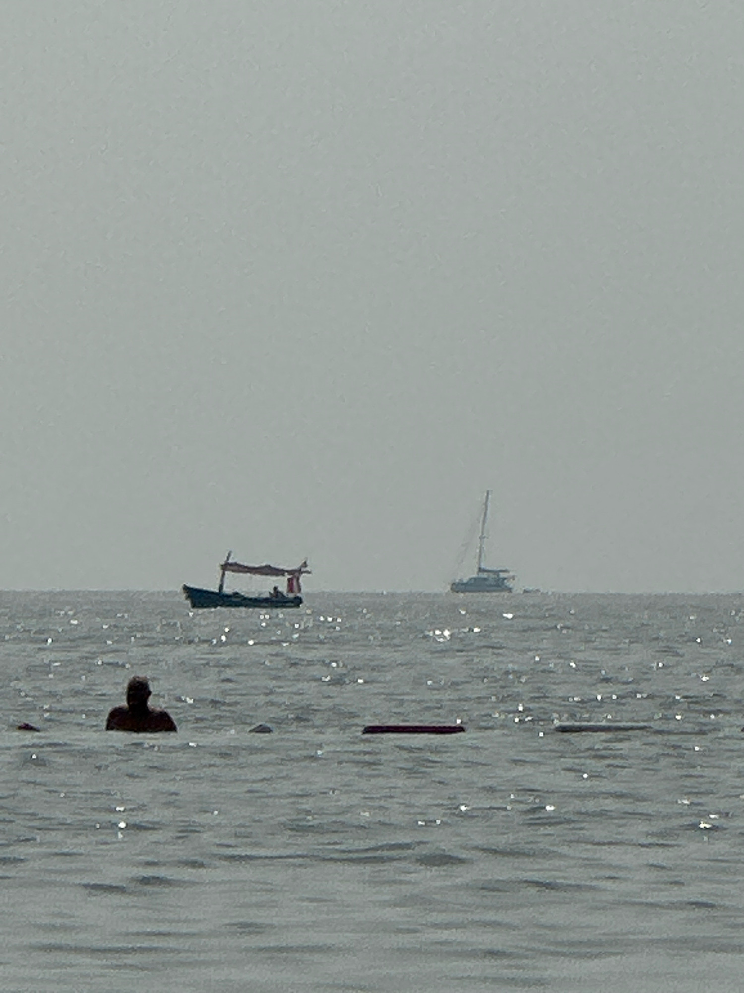 The image shows a body of water, likely the Adriatic Sea, with a hazy sky. In the foreground, a person is partially submerged in the water, with their head and shoulders visible. Further out in the water, there is a small boat with a canopy and at least one person inside. In the background, a sailboat with a tall mast is visible on the horizon. The water is choppy with sunlight reflecting off its surface, creating a shimmering effect. The overall scene suggests a calm day at sea, possibly near a coastal area.