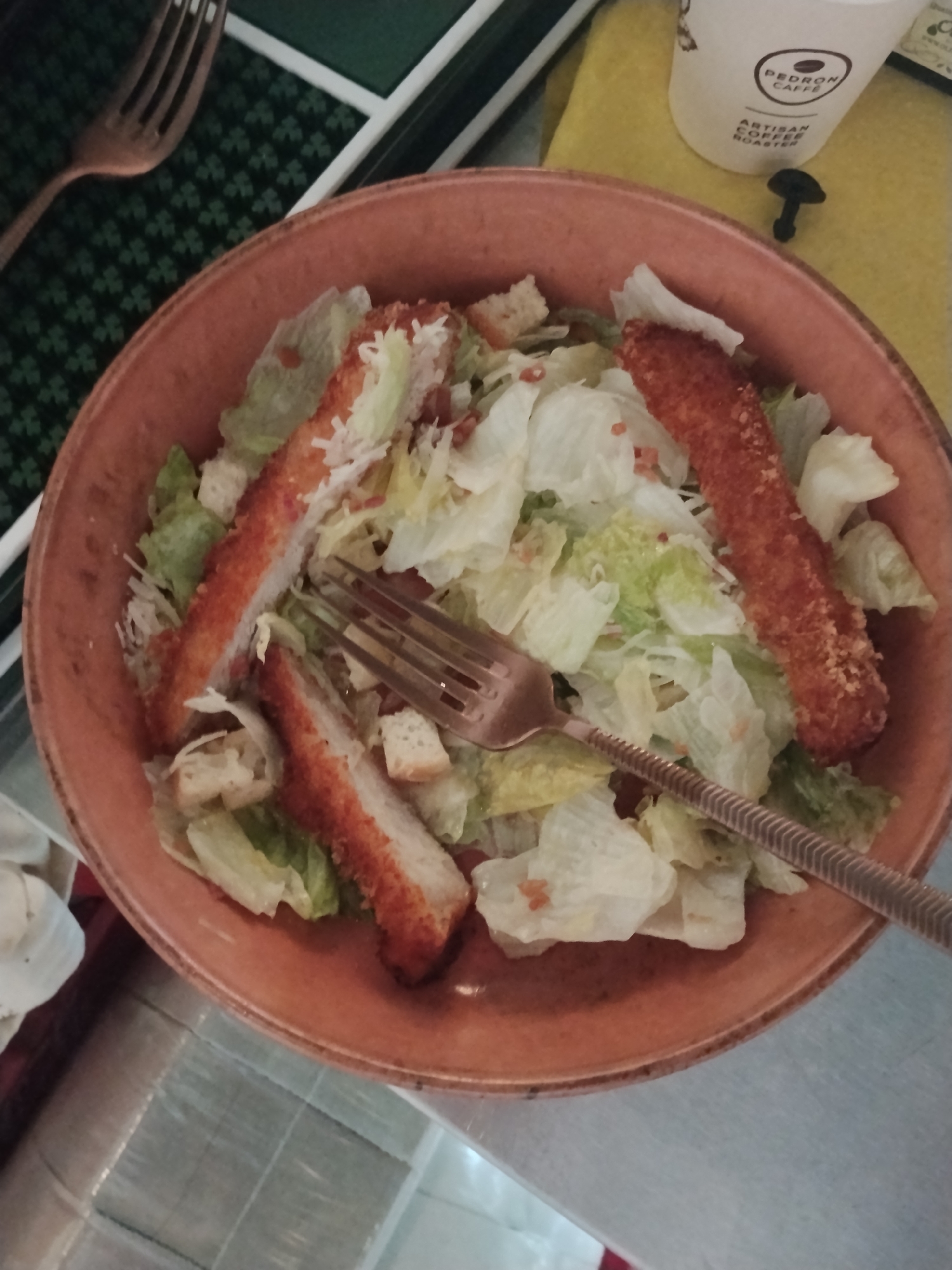 An overhead, close-up shot shows a Caesar salad in an orange bowl. The salad contains lettuce, croutons, and what appear to be breaded chicken strips. A fork is resting in the salad. To the right of the bowl is a white paper cup with a logo that says "PEDRON CAFFE" and "ARTISAN COFFEE ROASTER." A small, dark object, possibly a mushroom, is near the cup. The background is a yellow fabric placemat. To the left of the bowl, a fork rests on a dark green mat with a clover pattern. The scene appears to be set on a table with a metallic surface. There are no people visible.