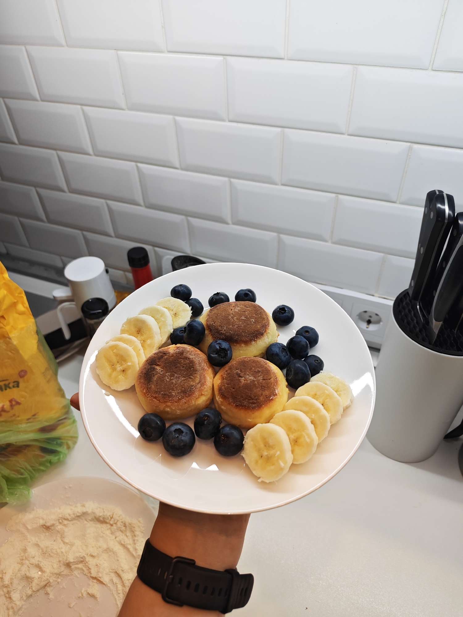 A person is holding a white plate of syrniki (cottage cheese pancakes) topped with blueberries and banana slices. The syrniki are golden brown. The plate is on a white countertop. In the background, there is a white tiled backsplash, a knife block with knives, and some kitchen appliances. To the left of the plate, there is a yellow bag of flour. In the foreground, there is spilled flour on another white plate. The scene is set in a kitchen, possibly in Bataysk, Russia, given the context provided.