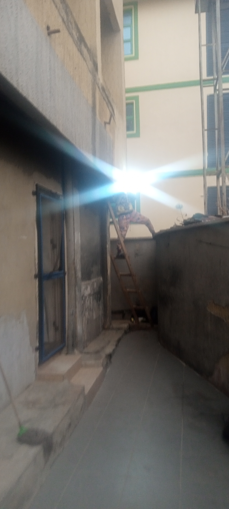 The image depicts an alleyway in what appears to be an urban environment in Kabala, Nigeria. The alleyway is flanked by two buildings; the one on the left is beige and has a blue-framed door, while the building on the right is unfinished concrete. The ground is covered in gray tile. A wooden ladder is positioned near the back of the alley, with an individual in patterned clothing visible on or near the top of the ladder. The lighting is poor, but there is a bright glare towards the back of the alley near the ladder. A broom is partially visible in the foreground to the left.