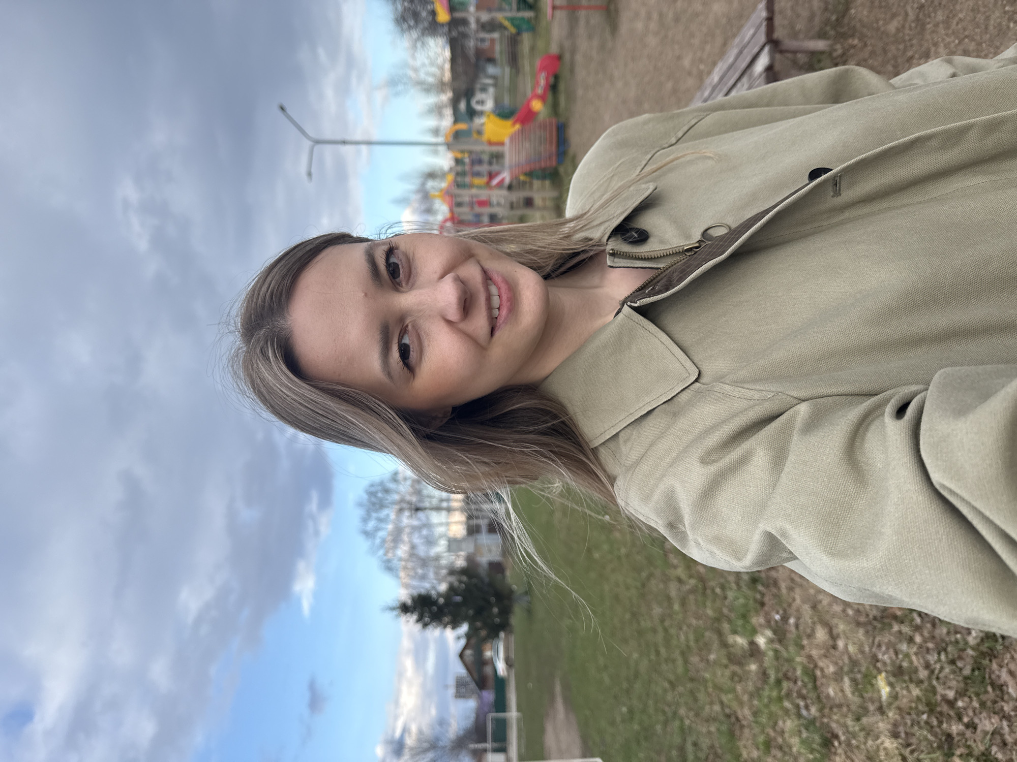 A casual, eye-level portrait features a young woman with light brown hair, smiling warmly and looking directly at the camera. She is wearing a light olive-green canvas jacket with a zippered and buttoned front closure, and a wide collar. Her head is slightly tilted, and her hair falls loosely over her shoulders.
The setting appears to be an outdoor recreational area or a park, possibly in a residential neighborhood in Chaikovsky, Russia. In the background to her left (from the viewer's perspective), a children's playground is visible, featuring colorful equipment like red slides, yellow climbing structures, and blue elements. A wooden park bench is partially seen behind her. Beyond the playground, scattered trees, a few distant houses or buildings, and a path or road indicate a suburban or town environment. A street light pole rises on the left side of the frame.
The sky above is partly cloudy with patches of clear blue, suggesting a daytime scene, possibly late afternoon, with soft, natural lighting. The ground is covered in green grass. There are no visible signs of specific activity other than the woman posing for the photograph. No text is visible in the image.