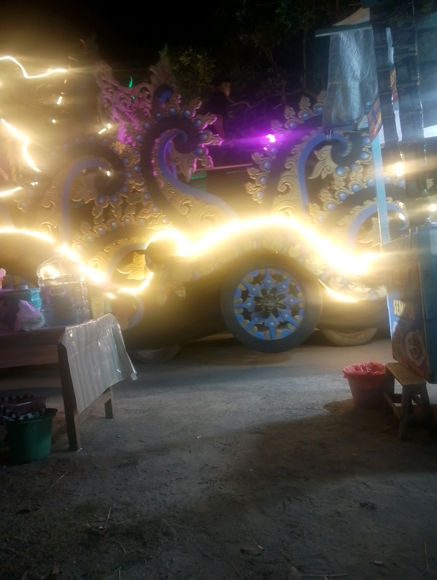 The image depicts a nighttime scene with a decorated vehicle, likely a parade float or a vendor's cart, illuminated by bright yellow and purple lights. The vehicle features ornate golden carvings with blue accents and a large blue-spoked wheel. In the foreground, a table covered with a translucent plastic sheet displays bottles of water and other items. To the right, a small wooden stool holds a red trash bin. The ground appears to be dirt or gravel, with some scattered debris. The overall impression is of a festive or commercial setup, possibly at an outdoor market or event in Ngebra, Indonesia. No people are clearly visible or identifiable in the frame.
