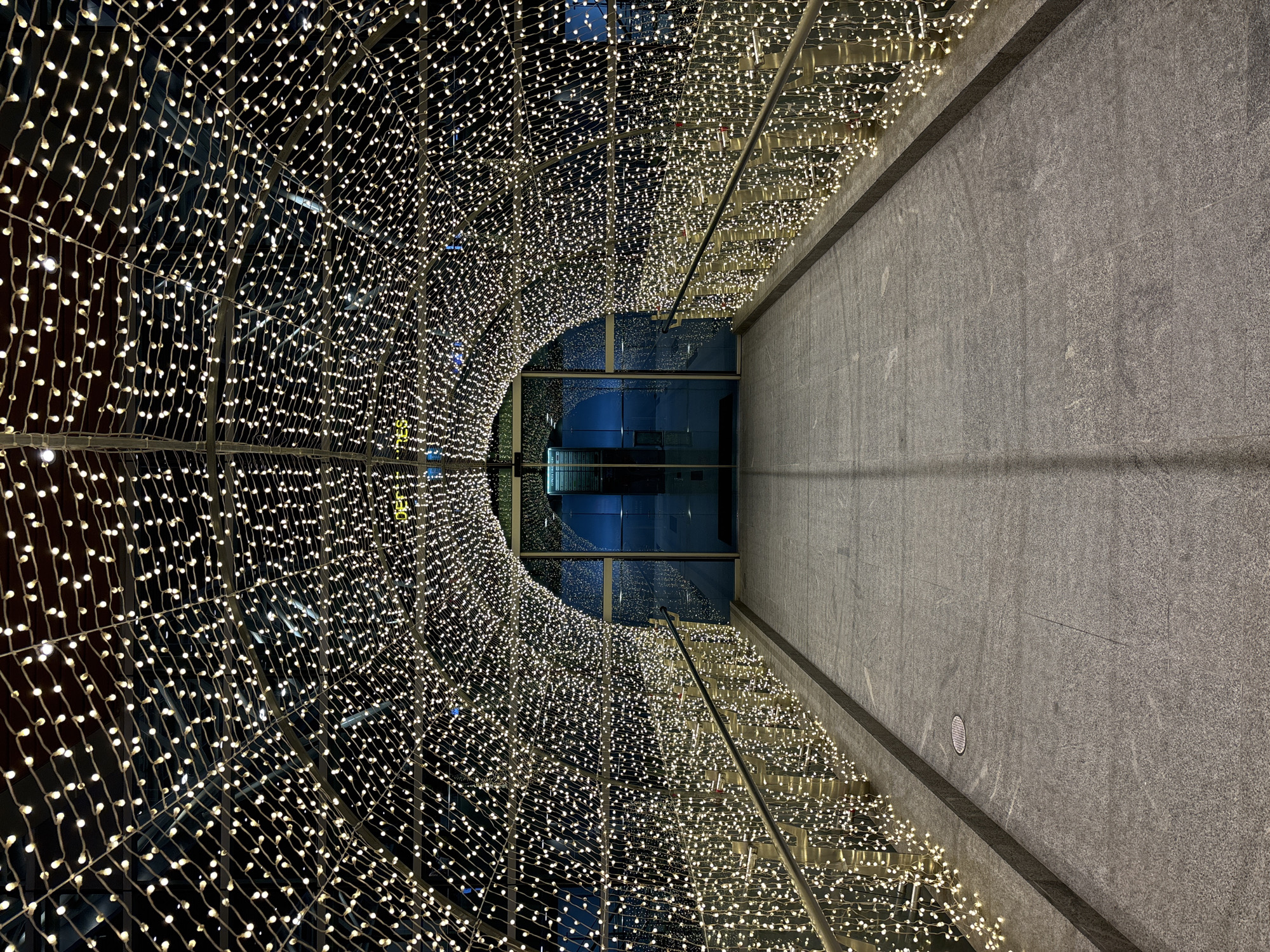 A low-angle, eye-level shot captures the interior of a light tunnel, likely in Đà Nẵng, Vietnam. The tunnel, constructed with metal framing, is densely covered in strings of small, warm-toned lights. The pathway of the tunnel is made of gray stone and is flanked by metal railings. In the distance, the tunnel opens to a building with large windows, reflecting blue light. The gray stone pathway extends out of the tunnel, suggesting it continues within the building. No people are visible.