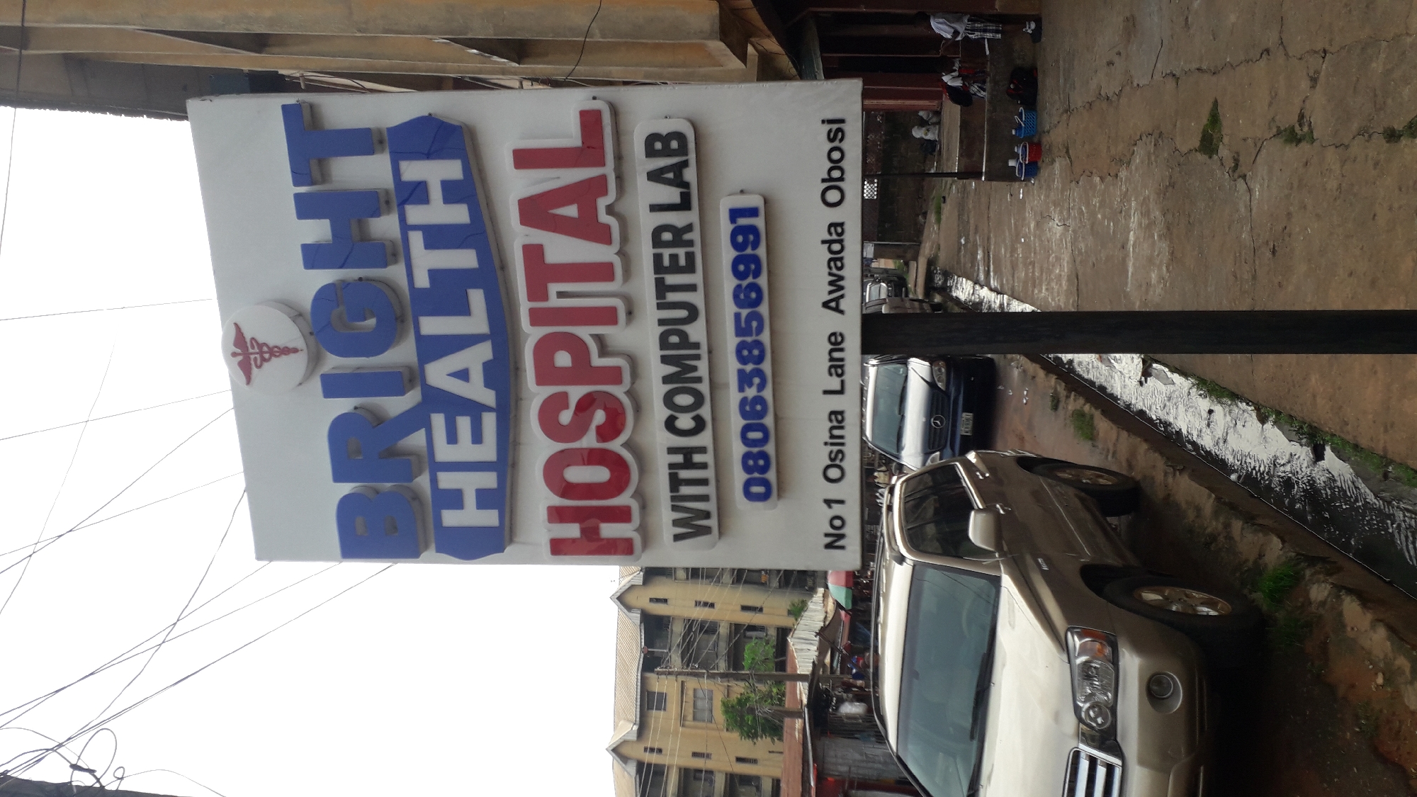 The image shows a sign for "Bright Health Hospital with Computer Lab," located at No. 1 Osina Lane, Awada Obosi, with the phone number 08063856991. The sign is blue and red on a white background. The caduceus symbol of medicine is visible on the upper left. Two cars, one silver and one dark colored, are parked nearby. The street appears wet, possibly from rain. A few people are visible in the background, possibly waiting by a building. The setting is a street scene in Onitsha, Nigeria. A multi-story building is visible behind the hospital sign.