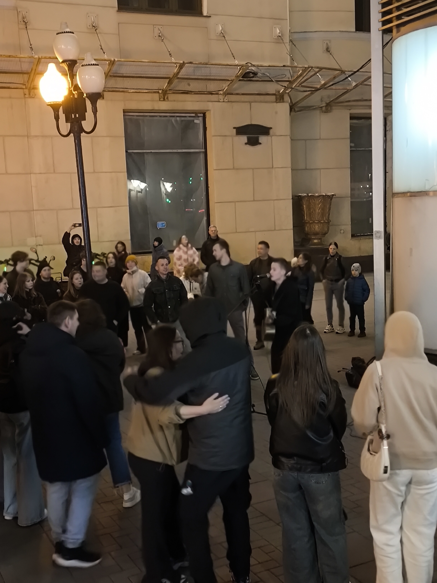 The image depicts a group of people gathered outside what appears to be a building in Moscow, Russia. The group consists of around 25 individuals of various ages, including children, adults, and possibly some teenagers. The majority of the people are standing, facing in a similar direction, possibly towards a performer or a central point of interest. Some of them are holding phones up, seemingly recording or taking pictures. Two individuals appear to be embracing in the foreground. The architecture of the building suggests an urban, European setting, with ornate details and a covered walkway. A streetlamp on the left illuminates the scene. The overall atmosphere seems relaxed and informal, possibly a street performance or gathering.