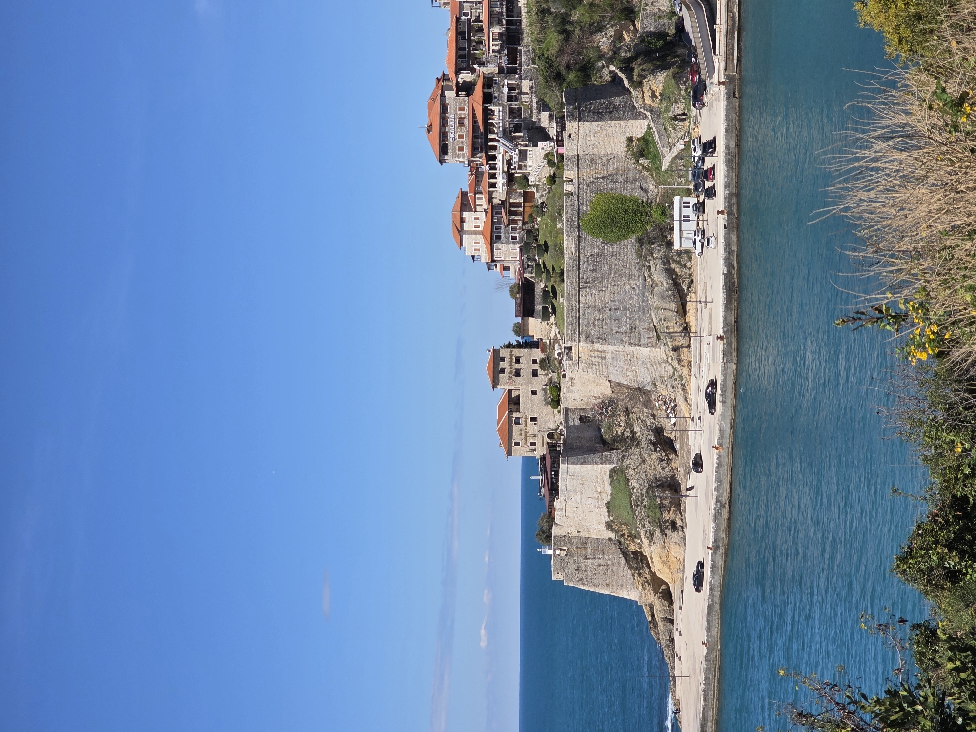 The image captures a sunny day in Ulcinj, Montenegro, focusing on a coastal scene. The composition is vertically oriented, with the blue sky dominating the left side and the coastline and water occupying the right. The coastline is a mixture of rocky cliffs and stone walls, supporting structures like buildings with red-tiled roofs. A road runs along the edge of the coast, featuring parked vehicles. The water is a clear blue, reflecting the sky. Vegetation is visible along the right edge of the frame. The image suggests a blend of urban development and natural landscape.