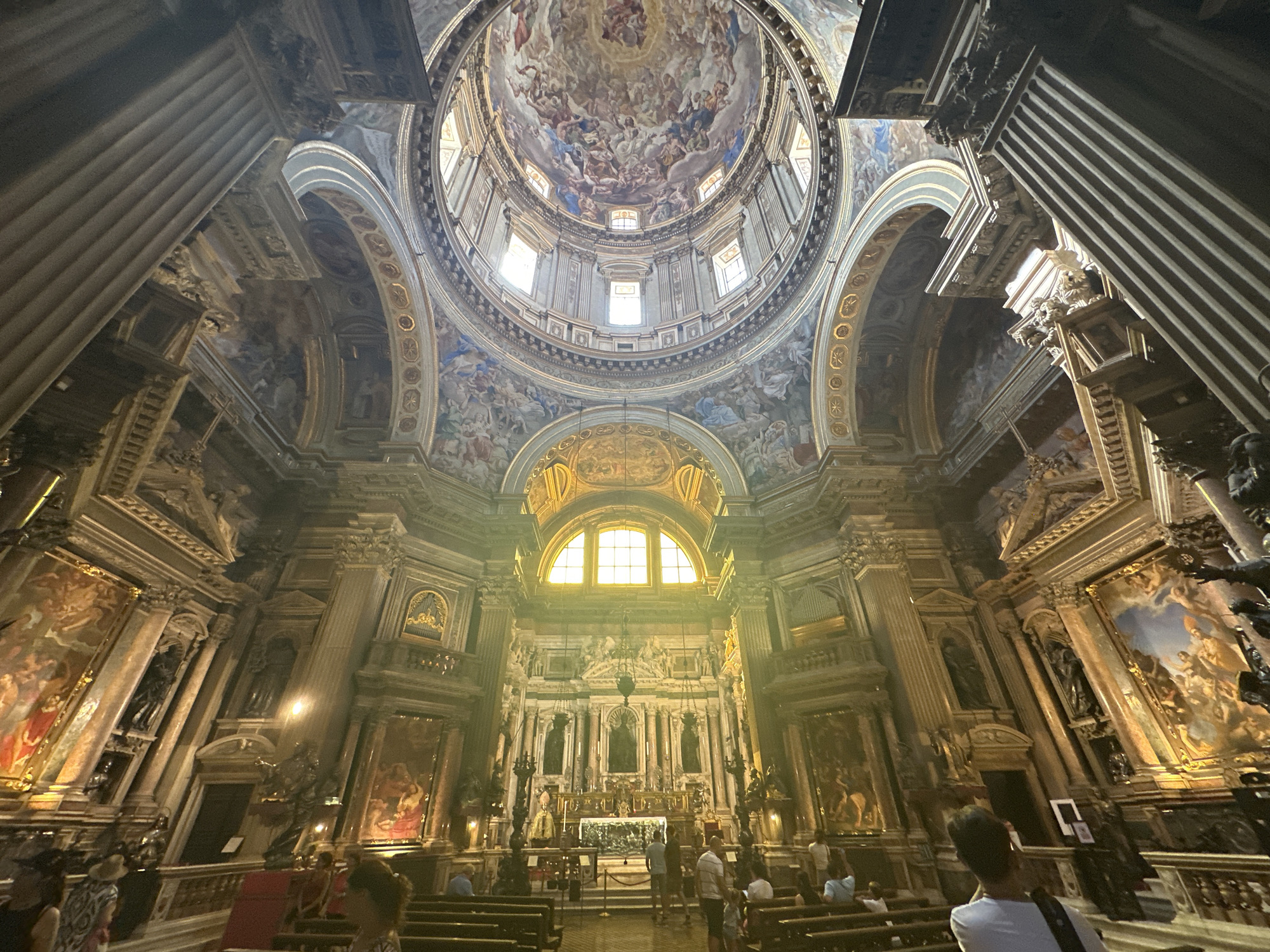 The image depicts the interior of the Church of the Gesù Nuovo in Naples, Italy, focusing on the nave looking towards the main altar and dome. The ornate Baroque architecture is evident, with richly decorated walls, columns, and the grand dome featuring extensive ceiling frescoes. Sunlight streams in from a large arched window above the altar, illuminating the scene and casting a golden glow. Several people are visible in the foreground and middle ground, some seated on pews, others standing and observing the architecture. The atmosphere appears to be one of quiet reverence and awe.