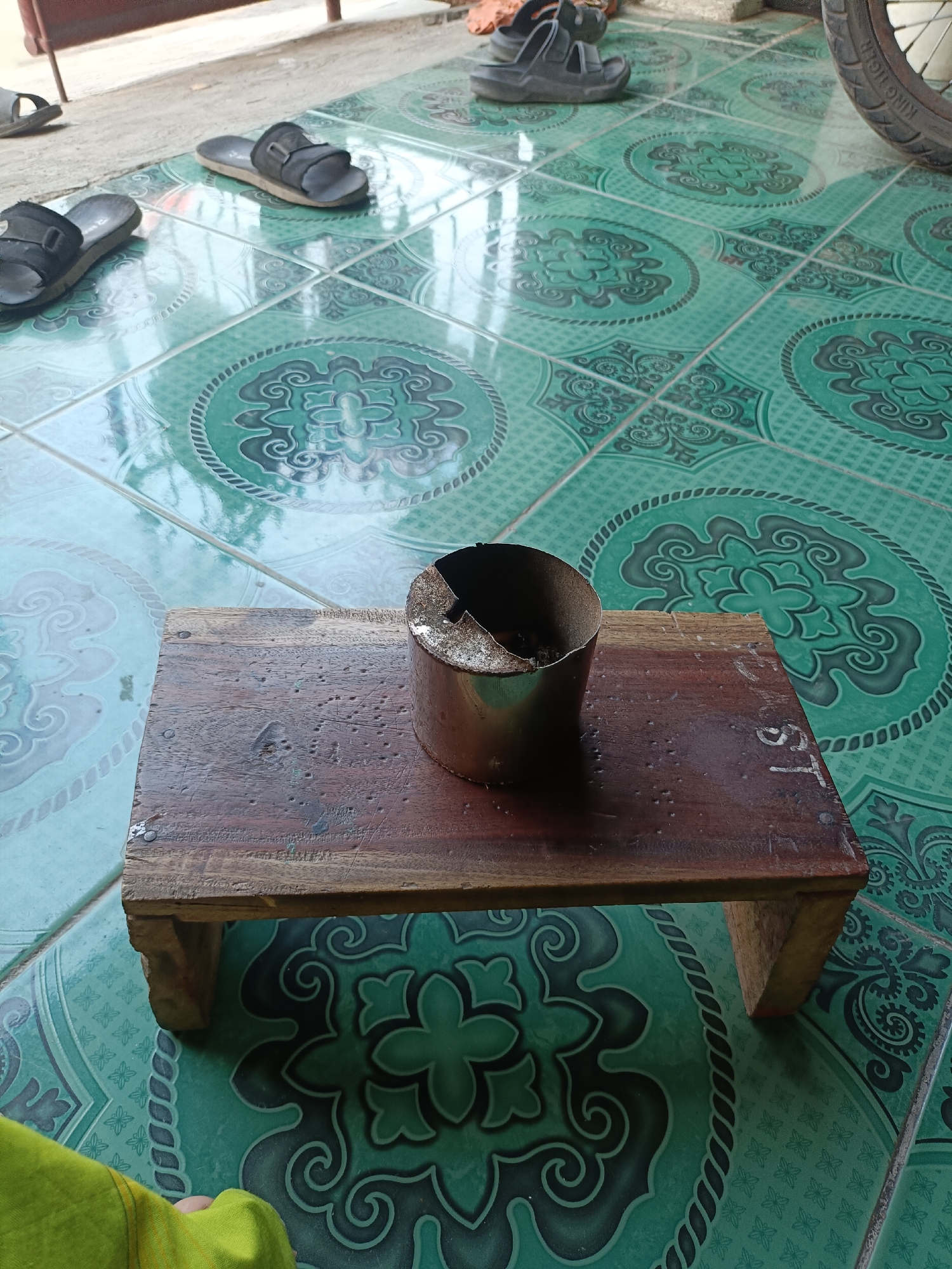 The image shows a close-up of a small wooden stool with a metal ashtray on top, resting on a green tiled floor. The tiles have a circular, floral pattern. Several pairs of black sandals are scattered around the floor, suggesting an outdoor or semi-outdoor area, possibly a patio or porch. In the background, part of a bicycle wheel is visible. The overall scene depicts a casual, lived-in space. The specific location in Telagamurni, Indonesia, is not visually evident from the image itself, but the environment suggests a residential or commercial setting common in such areas.