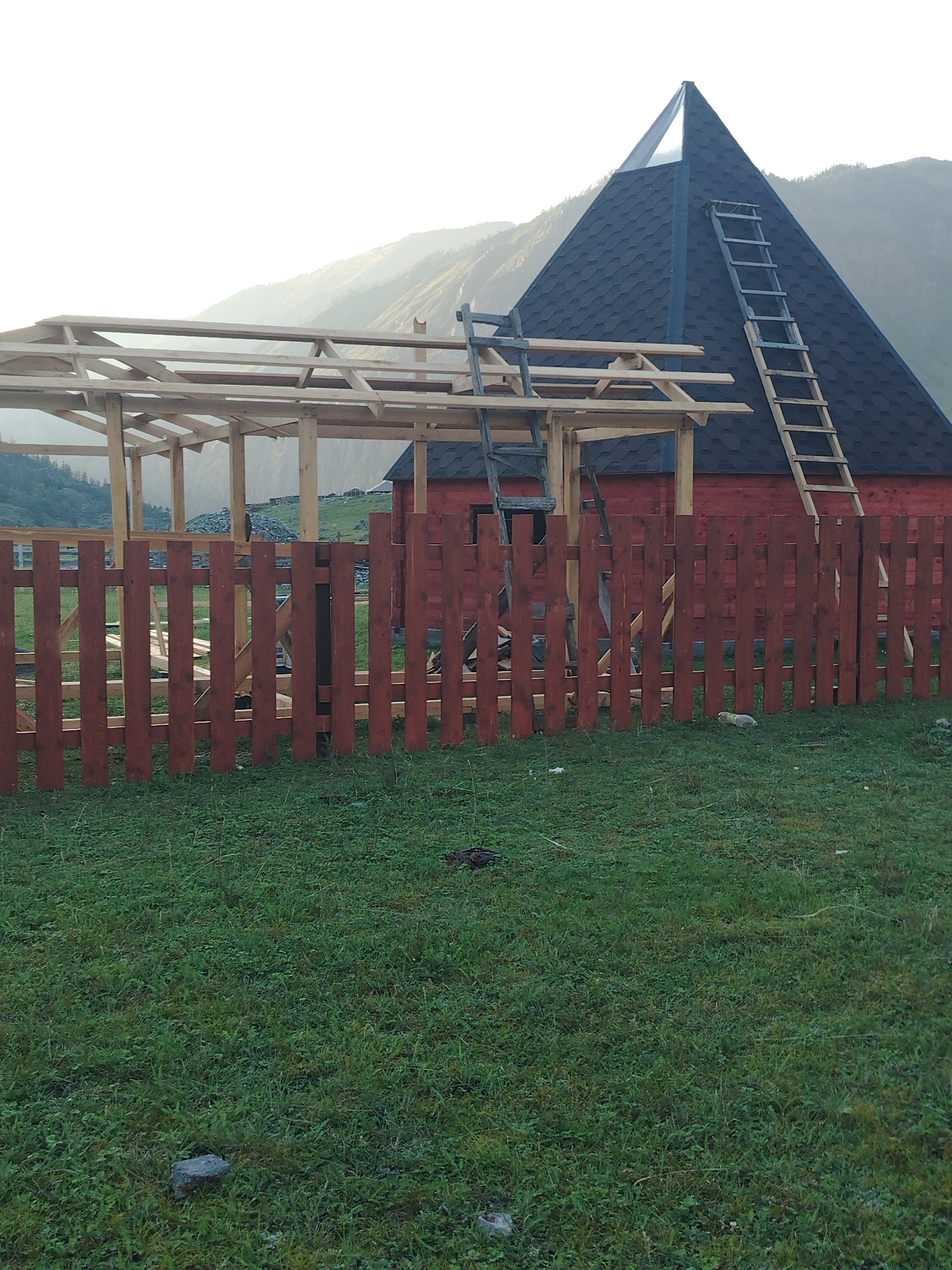The image shows a partially constructed wooden structure with a dark, multi-faceted roof, possibly resembling a pyramid shape, attached to a red building. A wooden ladder leans against the roof of the red building. In the foreground, a wooden fence made of vertical planks separates the building area from a grassy field. The background features rolling hills and mountains, with a hazy sky suggesting early morning or late afternoon light. The scene appears to be located in a rural or mountainous area, with no explicit indication of the city of Iodro, Russia, being present. The construction appears to be ongoing, with exposed wooden beams of the unfinished structure.