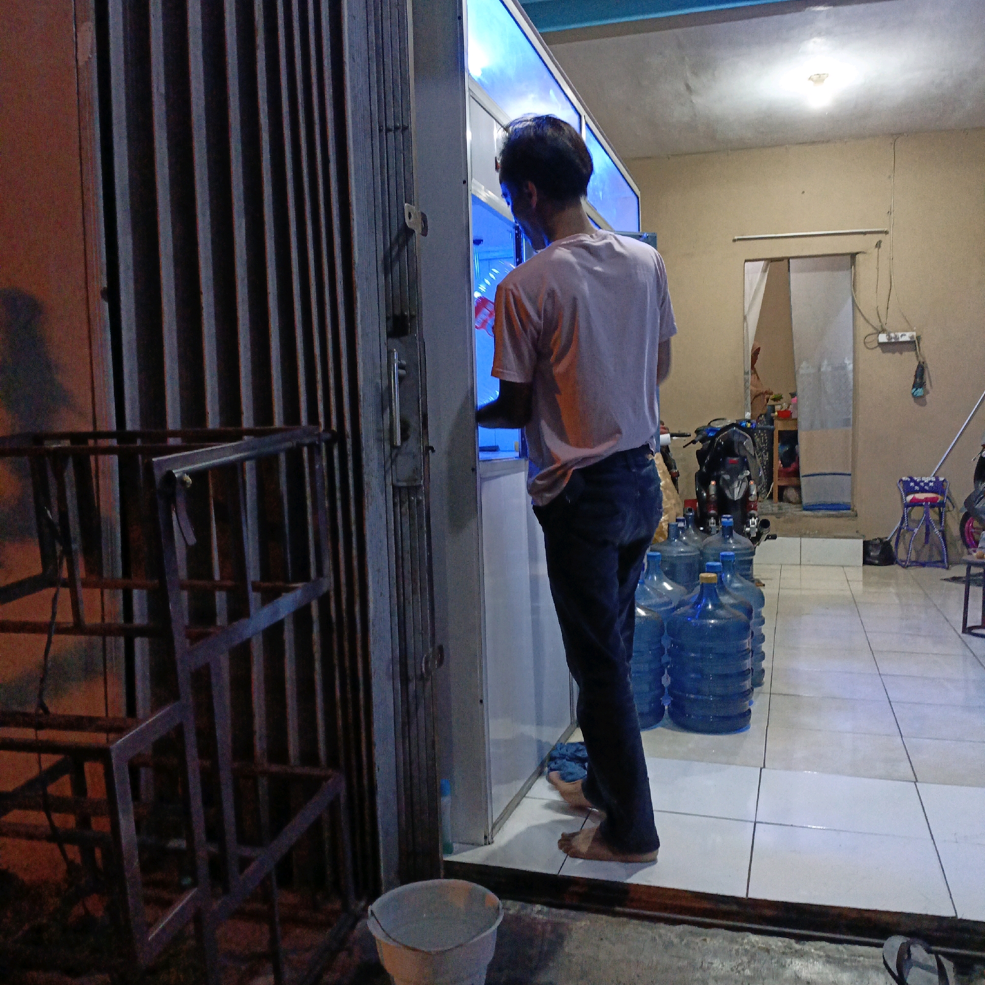 The image depicts an indoor scene at what appears to be a water refilling station located in the city of Salatiga, Indonesia. A young man, wearing a light-colored t-shirt and dark trousers, is standing with his back to the camera, interacting with a dispenser that is illuminated with blue light. In the foreground and to the right, several large, blue plastic water jugs are stacked. To the left of the man, a metal folding gate or barrier is visible. The environment is dimly lit, with the primary light sources appearing to be the dispenser and a ceiling light fixture. A motorcycle is parked in the background, further suggesting a commercial establishment. The floor is tiled. There's also a bucket in the lower foreground.