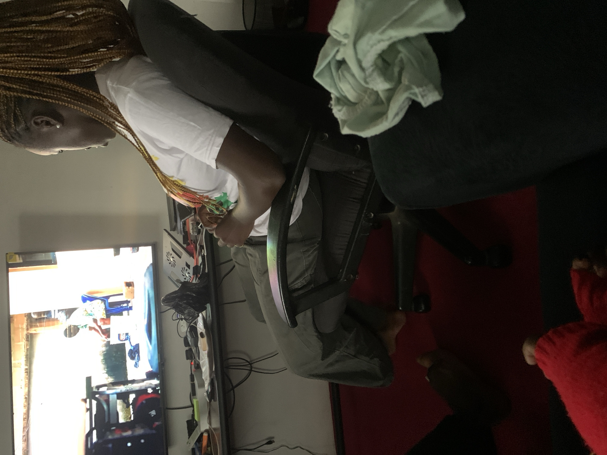 A side-profile medium shot shows a woman with long, braided blonde hair sitting in a dark grey office chair, facing a television. The woman is wearing a white t-shirt with a colorful graphic and dark green pants. Her left arm rests on the armrest of the chair, and her right hand is holding a light blue garment. The television screen shows a scene of a person in a brightly colored shirt sitting on a blue chair outdoors, near a white bucket. In the foreground, there are two pairs of feet visible, one wearing red socks and the other bare feet. The overall environment appears to be a living room, with a desk with electronic equipment and wires in the background. The floor is covered in a red carpet.