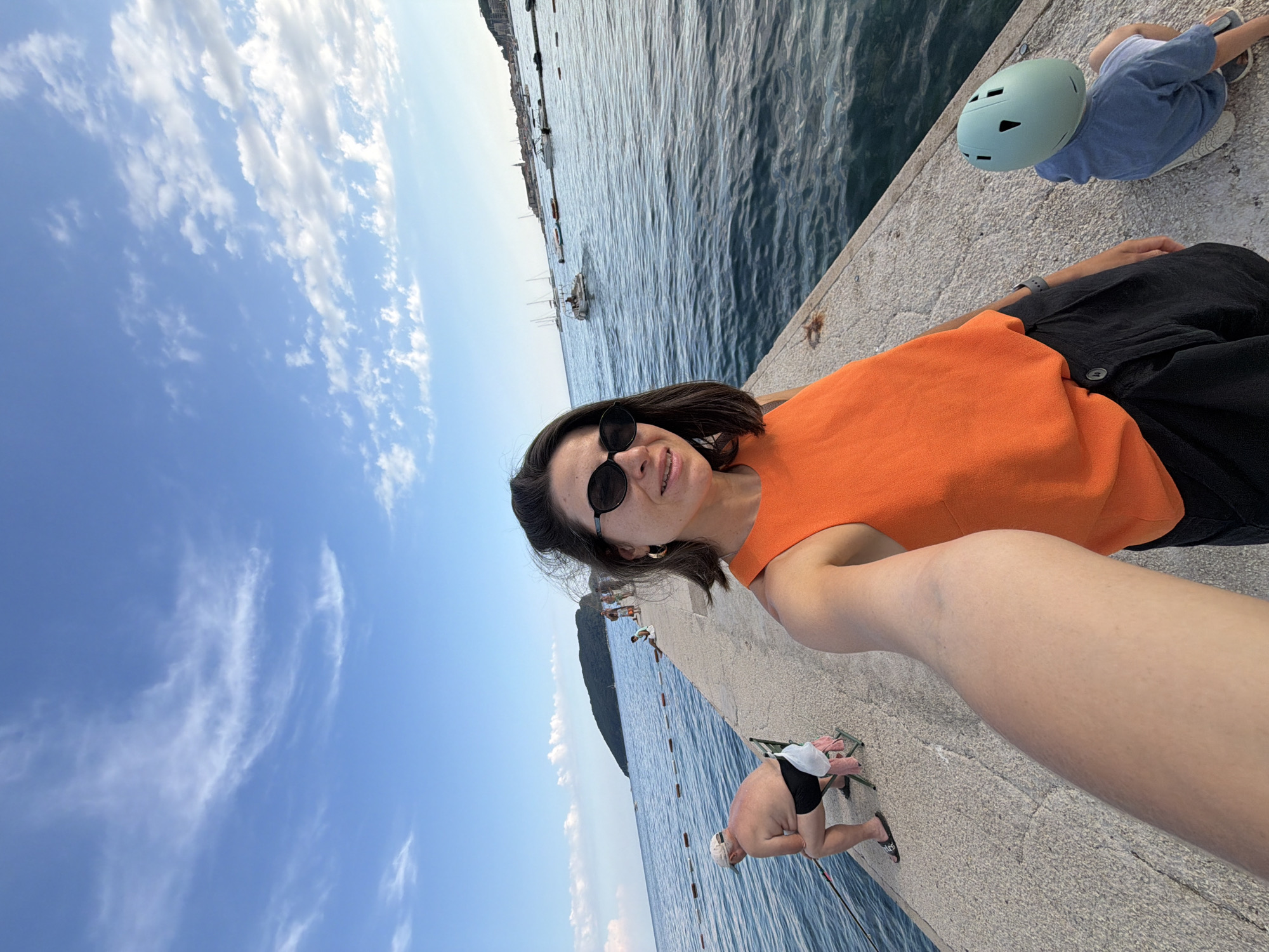 A woman is taking a selfie on a concrete pier in Budva, Montenegro. She is wearing an orange top and black shorts, with sunglasses and large hoop earrings. To her left, an elderly man is fishing, seated on a chair. To her right, a child wearing a light blue shirt and a light green helmet is sitting on the pier. In the background, the Adriatic Sea is visible, with boats and the coastline of Budva in the distance, under a blue sky with scattered clouds.
