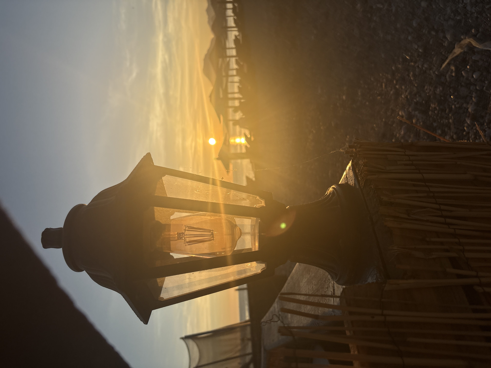A close-up, low-angle shot captures a vintage-style outdoor lamp in the foreground, its glass panels revealing an illuminated Edison bulb. The lamp is positioned on a textured wooden post wrapped with bamboo stalks. Beyond the lamp, a sun-drenched beach scene unfolds. The sun, low in the sky, casts a warm, golden glow across the horizon, creating a hazy, atmospheric effect. The sea is visible, with a few distant boats or figures on the water. The beach is dotted with lounge chairs and umbrellas, some of which are casting long shadows. Gentle waves can be seen lapping at the shore, and the pebbled surface of the beach is discernible. In the background, faint outlines of hills or mountains rise against the vibrant sky. The overall impression is one of a tranquil sunset at a coastal location, likely a beach resort. The specific location is confirmed to be Bar, Montenegro, suggesting a Mediterranean coastal setting.