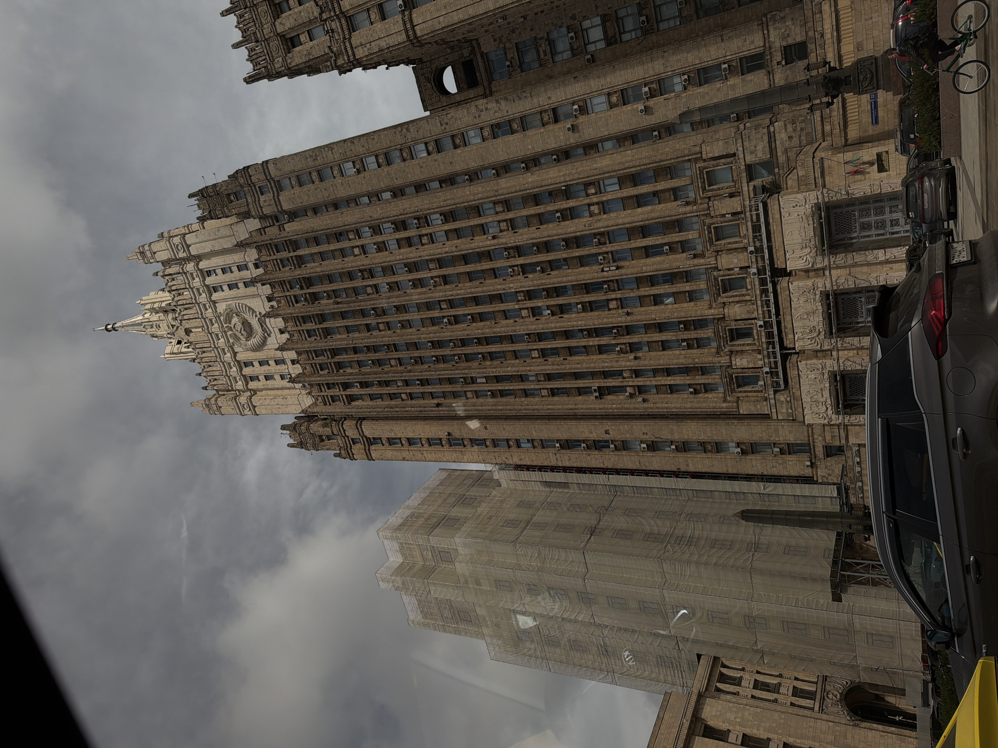 The image shows an upward view of several buildings in Moscow, Russia, under a cloudy sky. The most prominent building is a Stalinist skyscraper with a clock tower and spire, identified as the Ministry of Foreign Affairs. The building has multiple rows of windows, and some of the building is covered with construction tarps. A cyclist is visible near the building, along with several parked cars.