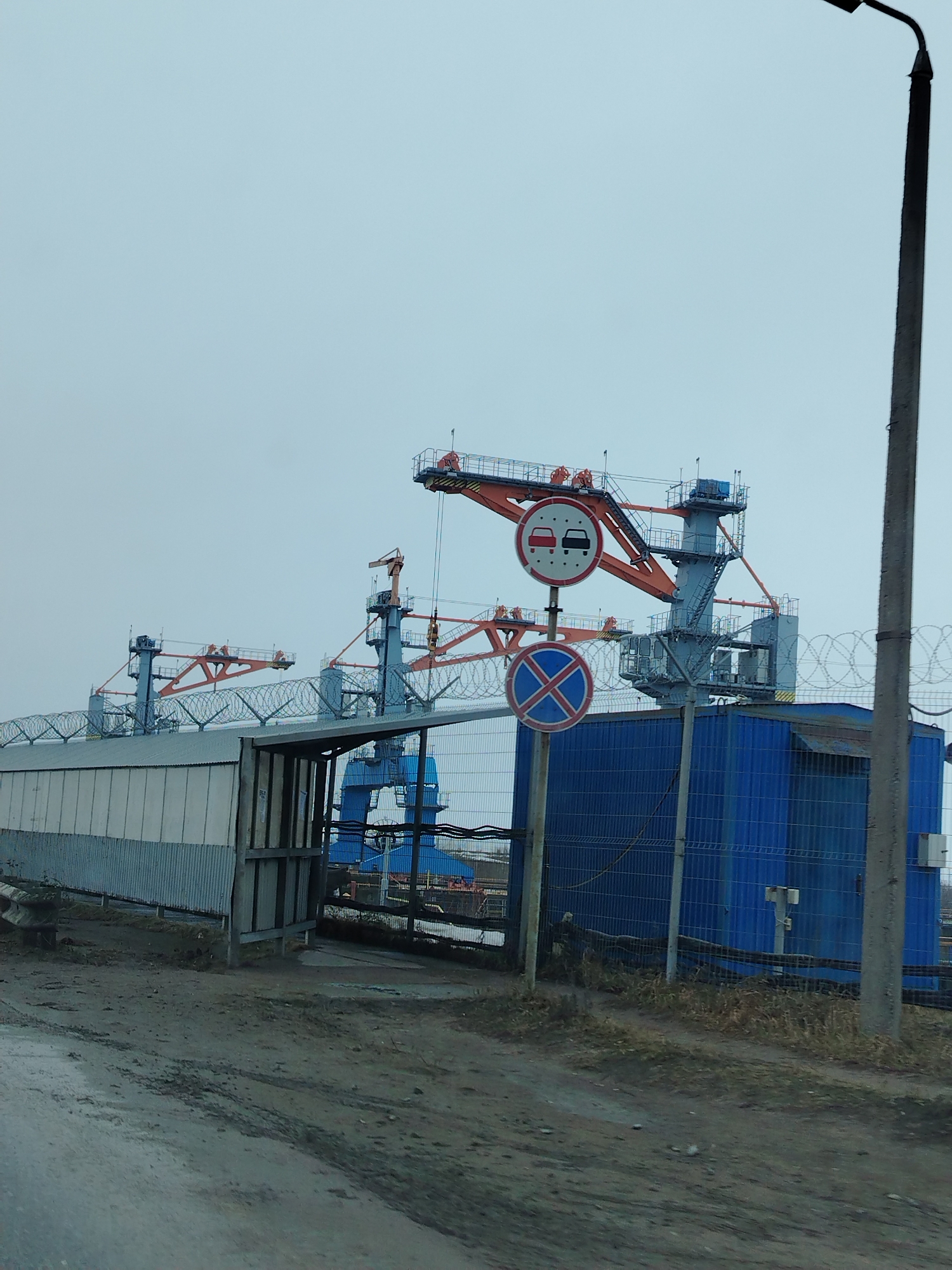 The scene depicts an industrial area near the city of Chaikovsky, Russia, identifiable by the architecture of the cranes and the presence of structures typical of industrial zones in the region. The sky is overcast, suggesting a dull or wintery day.
Several cranes are visible in the background. The cranes are predominantly gray and blue, with orange lattice work on their upper structure.
Foreground: A metal shed or fence of corrugated steel is to the left. A blue rectangular structure, potentially a building or large container, is to the right, enclosed by a chain-link fence topped with barbed wire.
In front of the blue structure, two traffic signs are mounted on a pole. The upper sign, a prohibition sign, depicts red cars overtaking black cars. The lower sign is a blue cross in a red circle.
The ground in the foreground is a muddy road with tire tracks. A gray light pole is on the right.