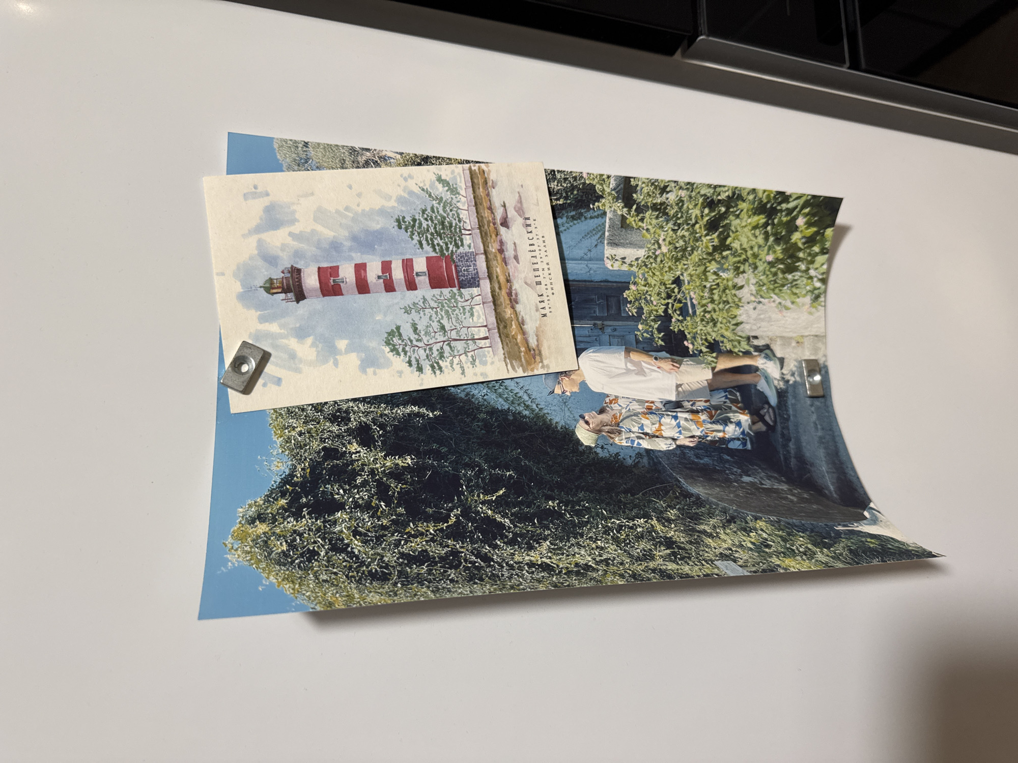 The image shows two postcards attached to a white surface by magnets. One postcard features a watercolor-style painting of a red and white striped lighthouse amidst sparse trees, with text in Russian identifying the artist as Mark Shedelevskiy and providing coordinates of 59° 59' 7 N 29° 36' 37 E, indicating a location near St. Petersburg, Russia, not Izhevsk. The second postcard shows a photograph of two people standing near what appears to be a stone archway, with lush greenery surrounding them. The image's background is a blurry white surface, potentially a refrigerator door or other appliance, with a dark, possibly metallic, upper edge visible at the top of the frame.