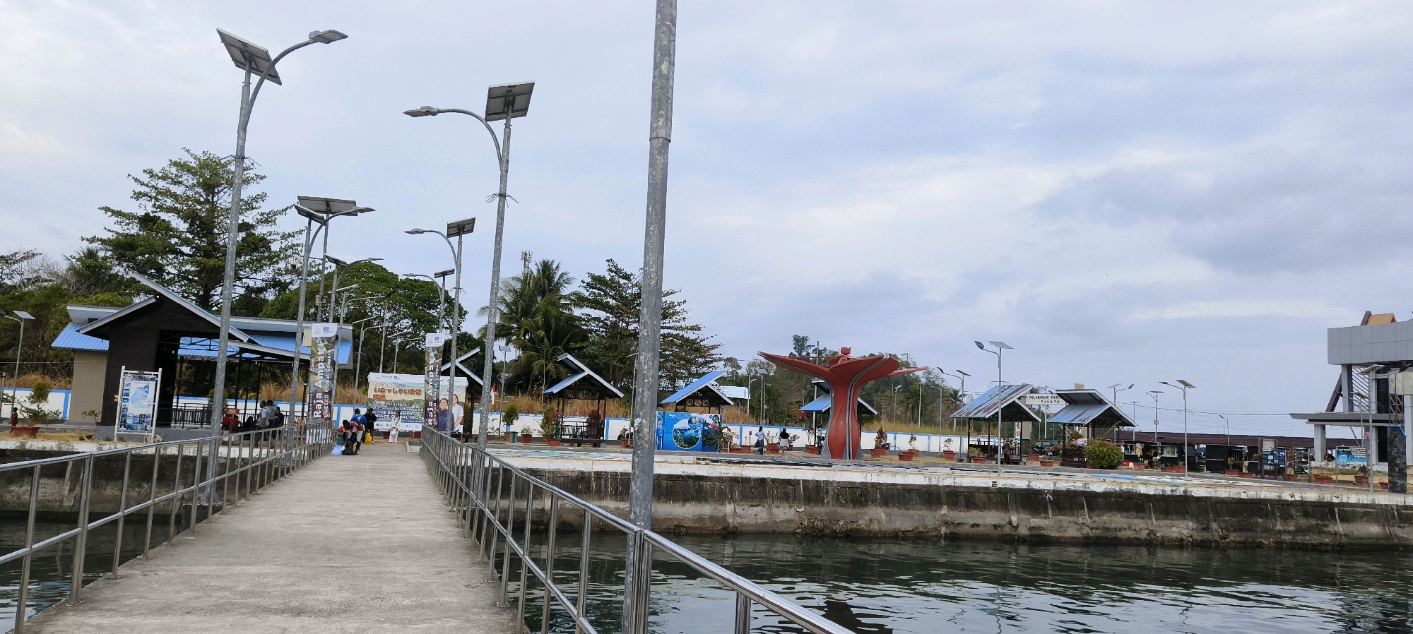 This is a photograph of a pier in Waisai, Indonesia, located in front of the Raja Ampat National Park. The pier is made of concrete and has metal railings on both sides. There are several solar-powered lamps lining the pier, as well as a few small covered shelters along the waterfront. In the background, there are trees and buildings, including what appears to be a visitor center or information booth. A few people are visible on the pier and in the background, some of whom appear to be waiting or sitting. The water is calm and reflects the cloudy sky. There is a large, red, abstract sculpture in the middle ground, resembling a stylized tree or flower. The overall impression is of a functional and somewhat developed tourist area.