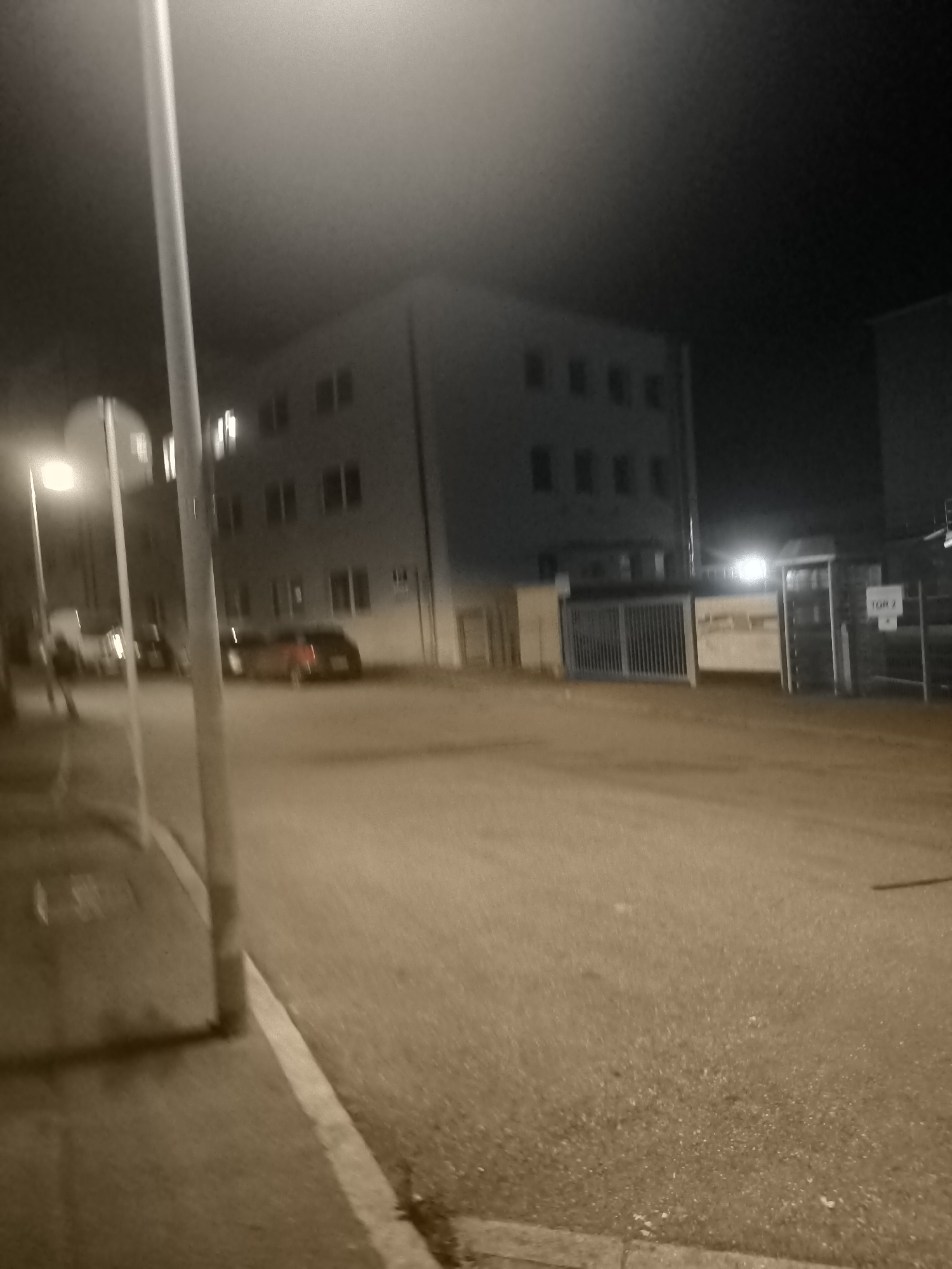 The image depicts an outdoor scene at night, likely in an urban environment. A street runs through the foreground, with a sidewalk on the left. On the right side of the street, there is a fenced area with a gate, and a sign that reads "TOR 2." Behind the fenced area, a building with multiple windows is visible. Across the street, another building with several floors and numerous windows is present, illuminated from within in some sections. Several cars are parked on the street, and at least one vehicle appears to be moving. A lamppost stands on the left side of the street, illuminating the area. The overall atmosphere is dimly lit, with the lighting primarily coming from artificial sources like streetlights and building interiors.