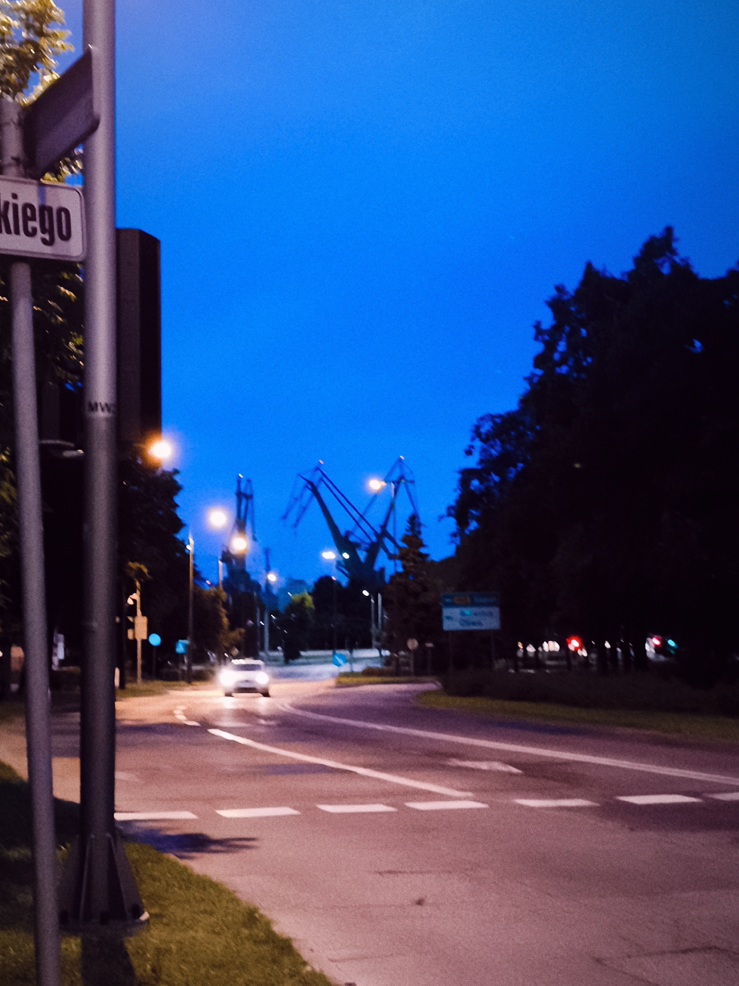 The image depicts an urban street scene at dusk or night, under a deep blue sky. In the foreground, on the left side of the frame, is a grey pole with a street sign partially visible, showing the word "skiego" which suggests a location in Gdańsk, Poland, likely relating to a street name. A car is visible in the middle ground, driving on a wet asphalt road with white lane markings. Further in the background, several large industrial cranes are silhouetted against the sky, indicative of a port or industrial area within Gdańsk. The overall atmosphere is dimly lit, with artificial lights from streetlamps and traffic lights casting a warm glow on the road. Trees line the side of the road, their foliage appearing dark and dense. The scene suggests a typical evening in an urban environment with maritime or industrial infrastructure.