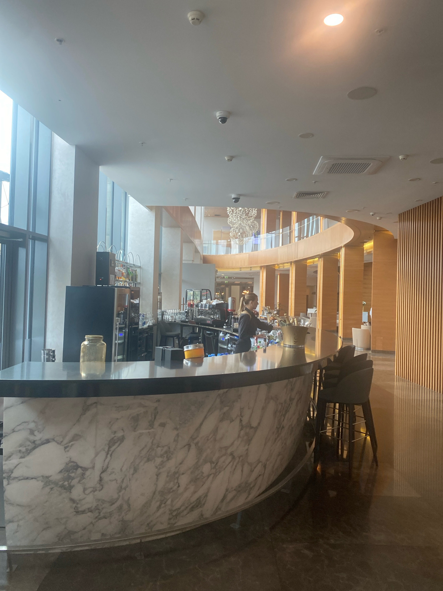 The image depicts the interior of a modern, upscale bar or lounge area, likely within a hotel or high-end establishment in Minsk, Belarus. A curved bar with a marble-patterned front is prominently featured in the foreground. Behind the bar, a female bartender with blonde hair tied back in a ponytail is visible, wearing a dark uniform. She appears to be attending to bottles and preparing drinks. Various bottles of alcohol and glassware are arranged on shelves behind the bar. In the center of the bar, there is an ice bucket, possibly containing champagne bottles. Several bar stools with dark upholstery are positioned around the bar.
The background reveals a spacious, contemporary architectural design with large windows on the left allowing natural light to enter. A distinctive chandelier hangs from the ceiling above a multi-level interior structure characterized by wooden paneling and curved elements. The flooring is a polished, dark material that reflects the ambient light. Multiple ceiling fixtures, including recessed lighting, air conditioning units, and security cameras, are visible. The overall atmosphere suggests a sophisticated and well-appointed venue.