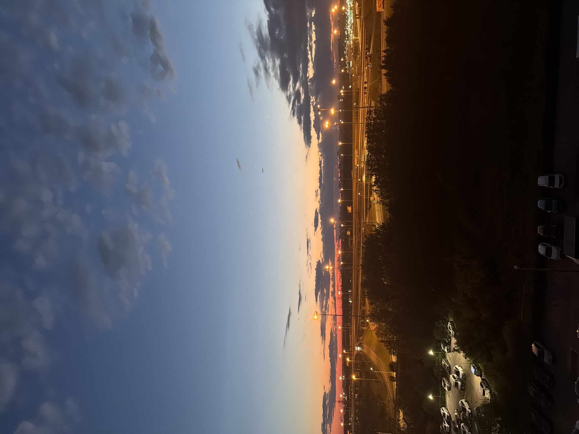 The image depicts an urban landscape at dusk, with a clear sky transitioning from blue to a gradient of orange and red near the horizon. A few scattered clouds are visible. In the midground, a complex highway interchange is illuminated by numerous streetlights. Trees and vegetation line the sides of the highway. In the foreground, a parking lot filled with cars is visible, some of which are illuminated by nearby streetlights. The overall scene is one of an active urban area transitioning into night.