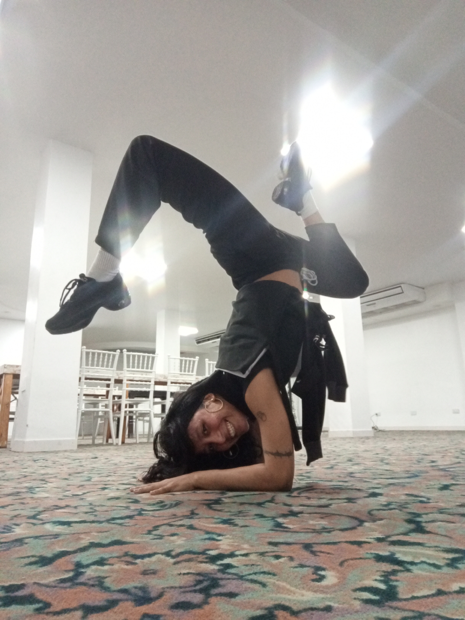 A female individual is captured in an inverted pose, likely a headstand or forearm stand variation, balancing on her forearms with her legs bent and arched above her head. She is looking directly at the camera with a wide, joyful smile. Her dark, long hair cascades around her face as she supports herself on a distinctively patterned carpet.
She is wearing a dark top, dark pants, white socks, and black athletic shoes. Visible accessories include large, gold-colored hoop earrings and a tattoo on her right forearm.
The setting appears to be a brightly lit indoor space, possibly a multi-purpose room or event hall, in Porlamar, Venezuela. The background features white walls and pillars, with several white-painted chairs and tables stacked or arranged in the distance, suggesting a cleared area for activities. A white wall-mounted air conditioning unit is also visible on the right. The ceiling is white, and a strong light source from above creates a prominent lens flare in the upper left and center of the image. The floor is covered with a patterned carpet featuring intricate swirls in shades of green, blue, pink, and beige.
The bright lighting and the subject's happy expression convey a positive and energetic mood. There is no visible text in the image.