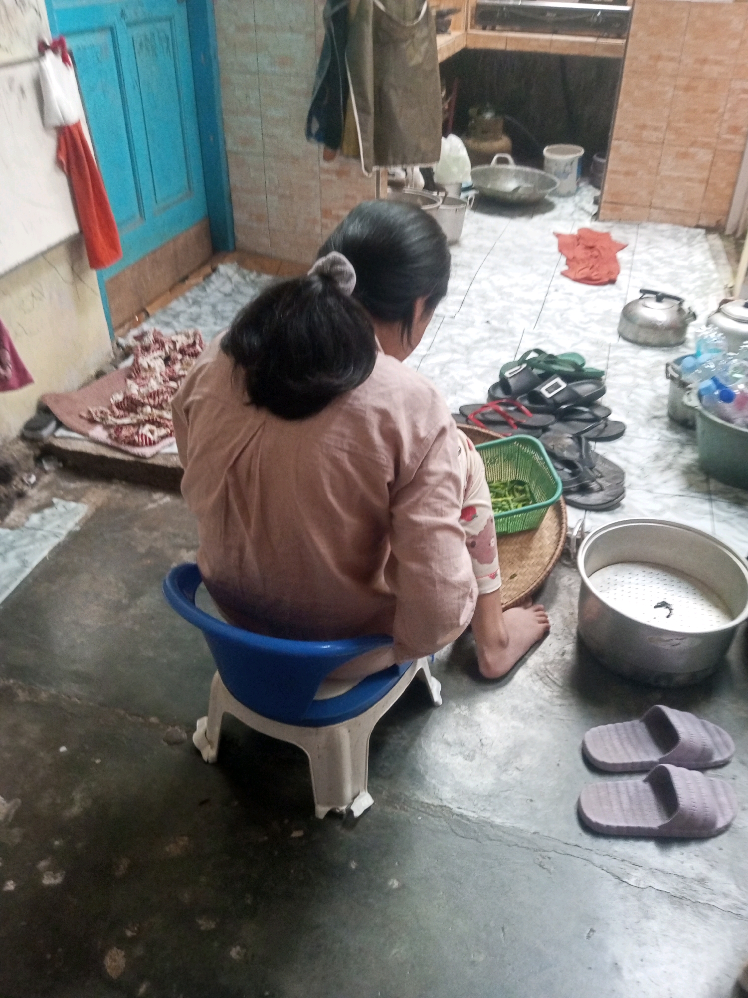 In Andongsili, Indonesia, a person is sitting on a blue plastic stool in what appears to be a home kitchen. The individual is facing away from the camera, their dark hair tied up. They are wearing a light-colored long-sleeved shirt and patterned pants. In front of them, on the floor, is a woven tray with a green basket containing food items, possibly vegetables. To their right, a stack of flip-flops is visible, along with a large metal pot with a steaming rack inside. In the background, kitchen cabinets and a blue door are present. The floor is concrete, and there are some red cloths and other items scattered around the room.