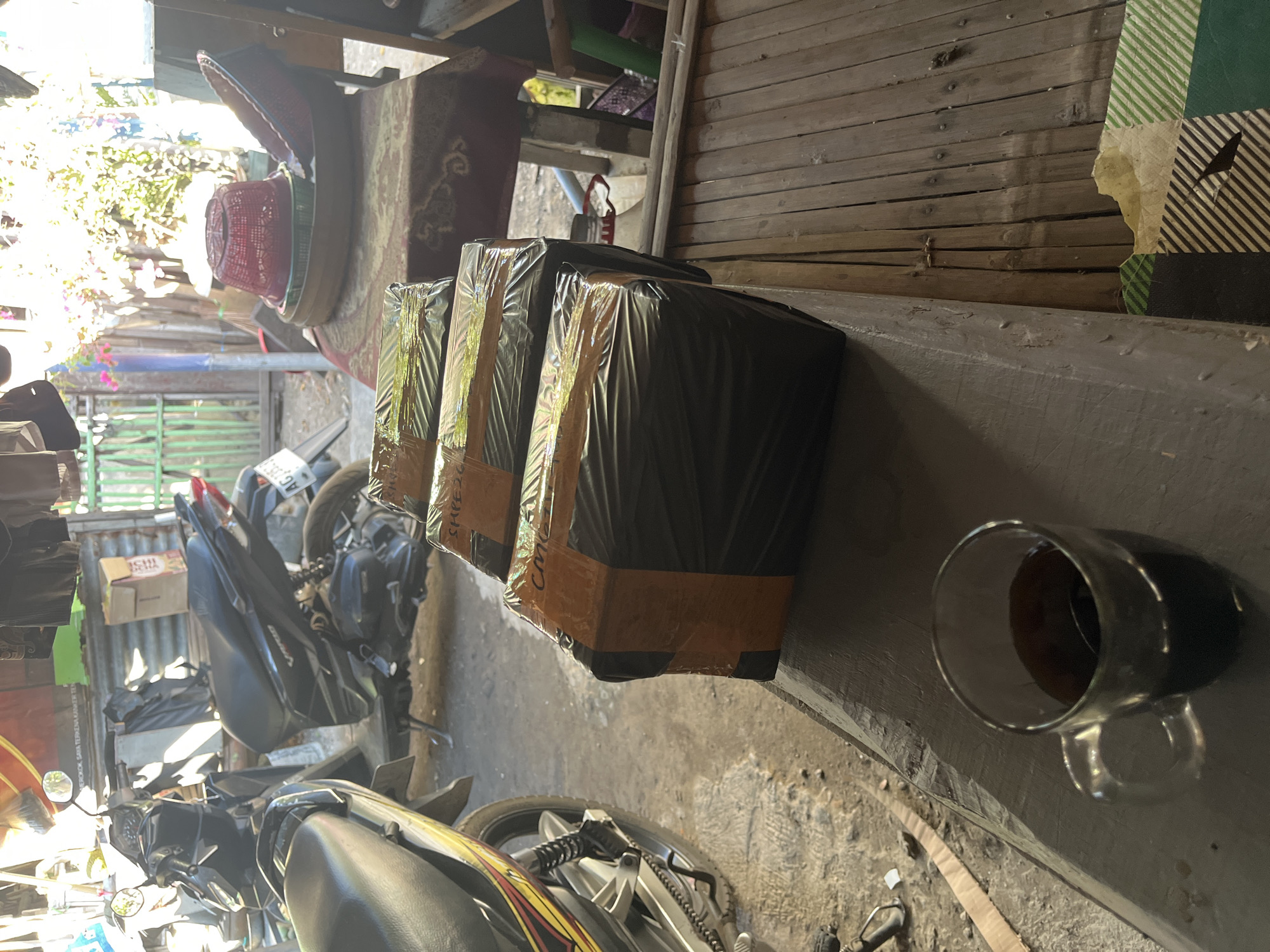 In Doroampel, Indonesia, the scene depicts an outdoor environment, possibly a yard or workshop, with a focus on stacked packages. Two motorcycles are parked in the background, suggesting a residential or small business setting. The packages, wrapped in black plastic and secured with orange tape, are arranged on a wooden surface in the foreground. A glass of dark liquid, likely coffee, sits next to the packages, indicating a moment of pause or waiting. The presence of a woven bamboo mat and some gardening materials in the background further contribute to the impression of an informal, functional space. The overall atmosphere is one of preparation or awaiting shipment.