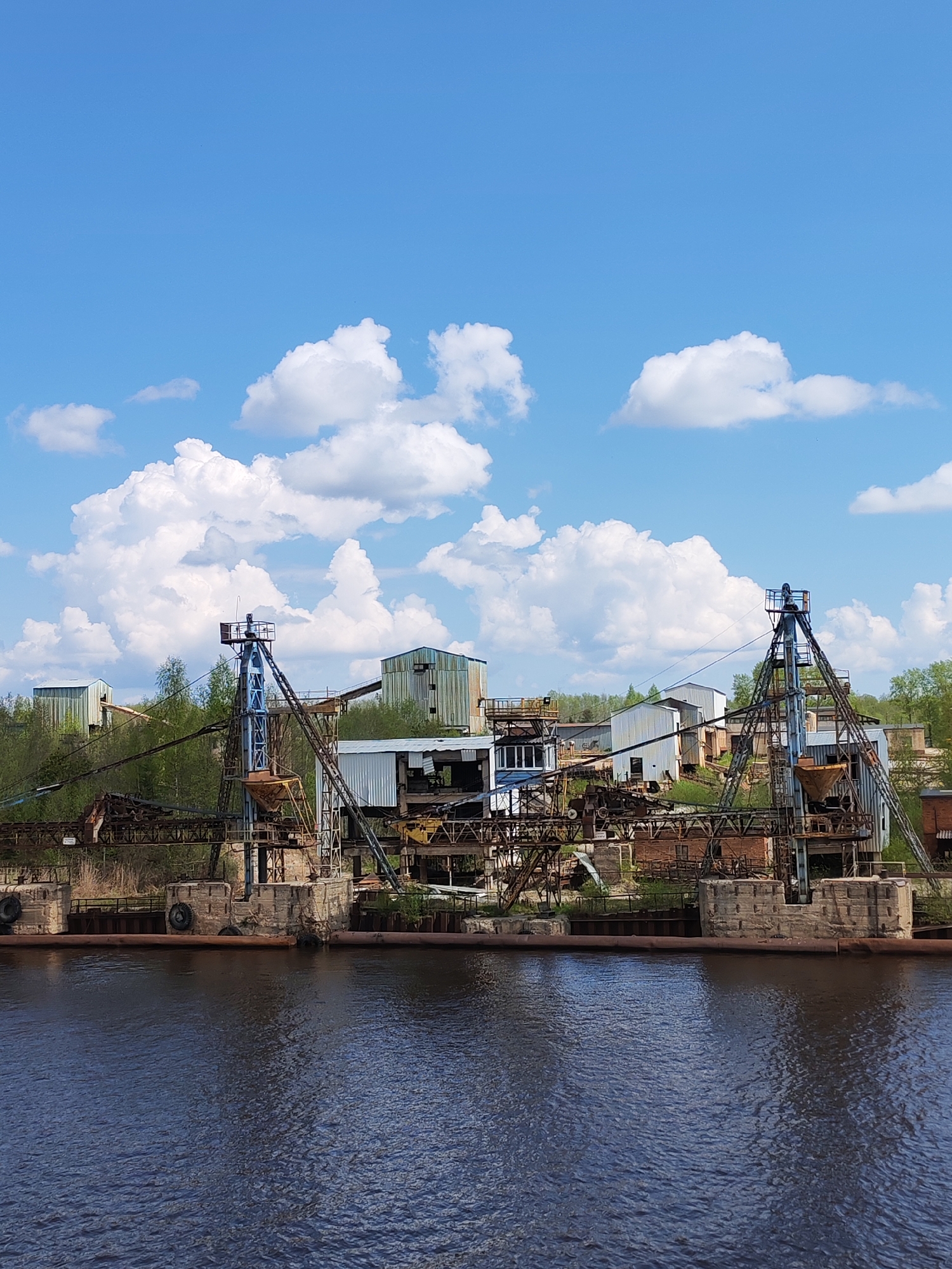 The image depicts a derelict industrial complex situated near a body of water. Two large, rusted cranes dominate the foreground, standing on weathered concrete platforms reinforced with tires. These cranes are connected by a network of rusted metal supports. Behind the cranes are a collection of dilapidated industrial buildings with corrugated metal siding and various structural components exposed. The buildings are interspersed with patches of green vegetation. The water is dark and rippled, reflecting the overcast sky. Above, the sky is blue with scattered white clouds. The complex is located in the city of Ivanov Bor, Russia.