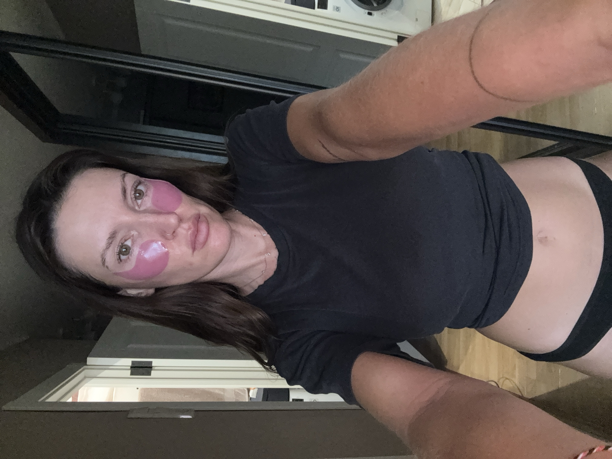 A woman with brown hair is standing in a room. She is wearing a black t-shirt and black underwear. She has pink under-eye patches on. Her arms are extended forward, and she is holding a phone, taking a selfie. In the background, there is a door, a mirror, and a washing machine. The floor is made of wood planks. There are no visible indications of the image being located in Moscow, Russia.