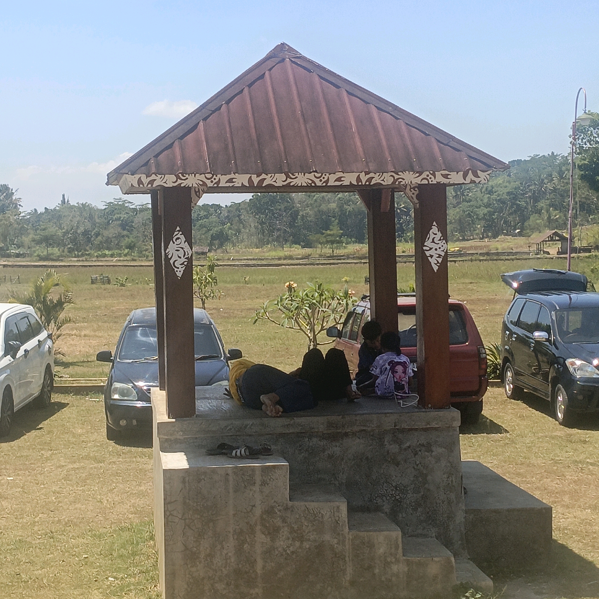 In the city of Krajan Lor, Indonesia, a small, open-air pavilion with a dark red, corrugated metal roof stands in a grassy, outdoor area. The pavilion has four dark wooden posts, two of which are adorned with white, decorative patterns resembling stylized foliage. Underneath the pavilion's roof, three individuals are present. One person, wearing a yellow shirt and dark pants, is lying down, facing away from the viewer, with their feet visible. Two children, one wearing a purple shirt with a graphic print and the other in a dark blue shirt, are sitting. A backpack with a colorful pattern is placed near the children.
Outside the pavilion, several vehicles are parked on the grass. To the left, a white car is partially visible. In front of the pavilion, a black car is parked. To the right of the pavilion, a red car is visible, and a black SUV with its rear hatch open and a roof carrier is parked further to the right.
The ground around the pavilion is covered in dry grass. In the background, beyond a line of trees and some agricultural fields, a clear blue sky with a few scattered clouds dominates the scene. The overall impression is a relaxed outdoor gathering in a semi-rural setting.