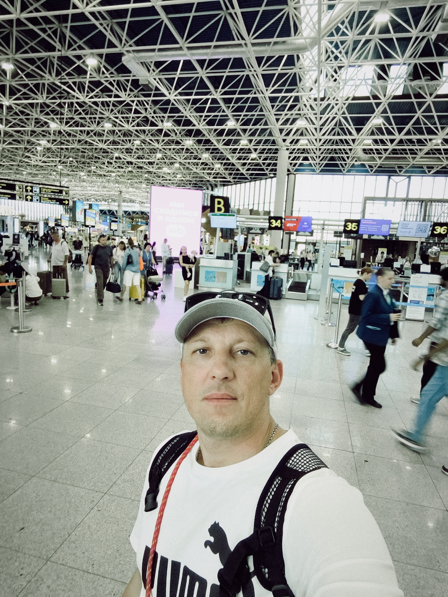 The image shows a selfie taken in an airport terminal. A man wearing a white Puma t-shirt and a gray baseball cap is in the foreground, looking directly at the camera. He has a backpack on. Behind him, the airport is busy with people and luggage. Several check-in counters are visible, with signs indicating gate numbers like "34", "35", and "36". A large pink digital display board with text in Russian is also prominent. The architecture of the airport features a complex, modern ceiling structure with exposed metal beams and lighting. The floor is tiled and reflective, with the lights from the ceiling creating reflections. People are seen walking with luggage, suggesting travel. The overall atmosphere appears to be that of a typical, bustling airport environment. The Russian text on the large display board translates to "We meet in one" and "wb", likely related to an airline or travel service. The location context provided is Sochi, Russia. The time of day is not explicitly indicated, but the lighting suggests it could be daytime. No specific emotions are overtly displayed by the individuals in the background.