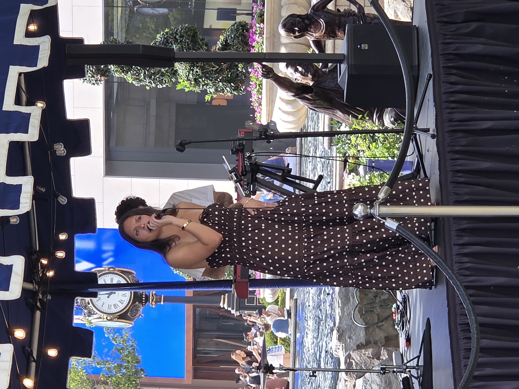 A woman in a brown and white polka dot strapless dress sings into a microphone on a stage. She is wearing a gold bracelet. Behind her, a person wearing a white shirt and a dark curly wig stands. A black speaker is to the right of the stage, and a black microphone stand is to the left. In the background, a large clock with Roman numerals is visible, and a fountain with clear water is also in the background. The overall scene appears to be an outdoor performance stage in Glendale, United States of America.