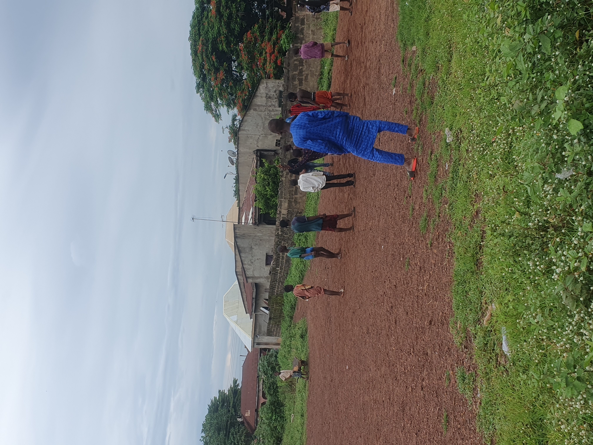 The image captures an outdoor scene in Idah, Nigeria, under an overcast sky. The foreground consists of a strip of green grass interspersed with small white flowers and some discarded refuse. This transitions into a wide expanse of reddish-brown, unpaved earth occupying the midground. The background features various single-story and two-story residential or commercial buildings constructed from concrete blocks, displaying diverse roof materials including corrugated metal and flat designs. A large tree with distinctive red-orange blossoms is prominent on the right side of the background, and multiple satellite dishes are affixed to rooftops.
Eleven individuals, predominantly appearing to be male youths, are distributed across the unpaved area and the adjacent grassy patch. In the immediate foreground, a young male stands with his back to the viewer, clad in a bright blue traditional tunic and matching trousers, complemented by orange and dark sandals. He is oriented towards the other individuals.
To the left, a young male in a light-colored shirt and dark trousers stands next to a yellow wheelbarrow on a grassy section. On the reddish-brown ground, a group of youths is scattered. Their attire includes various shirts, a patterned tunic, bare torsos, and shorts in colors such as green, orange, purple, and red. Their postures suggest an informal gathering or casual interaction within the open space. The sky is uniformly grey with visible cloud formations.