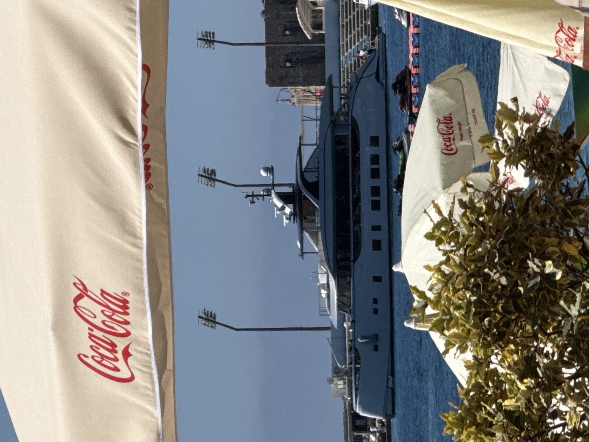 The image shows a large yacht docked in a harbor. The yacht is gray and has multiple levels with many windows. In the foreground, there are several white umbrellas with the Coca-Cola logo visible on them. A green tree with dense foliage is also present in the foreground, partially obscuring the yacht and umbrellas. In the background, a stone structure, possibly a building or fortification, can be seen. The sky is clear and blue, suggesting a sunny day. The scene appears to be a waterfront setting, likely a marina or harbor in a coastal town. The presence of umbrellas suggests an outdoor seating area, perhaps for a cafe or restaurant. No people are clearly visible in the image, though there are hints of figures on the yacht. The overall mood is relaxed and scenic. The visible text is "Coca-Cola" on the umbrellas. The scene suggests a daytime setting, likely warm weather given the clear sky and outdoor umbrellas. The location is identified as Budva, Montenegro.
