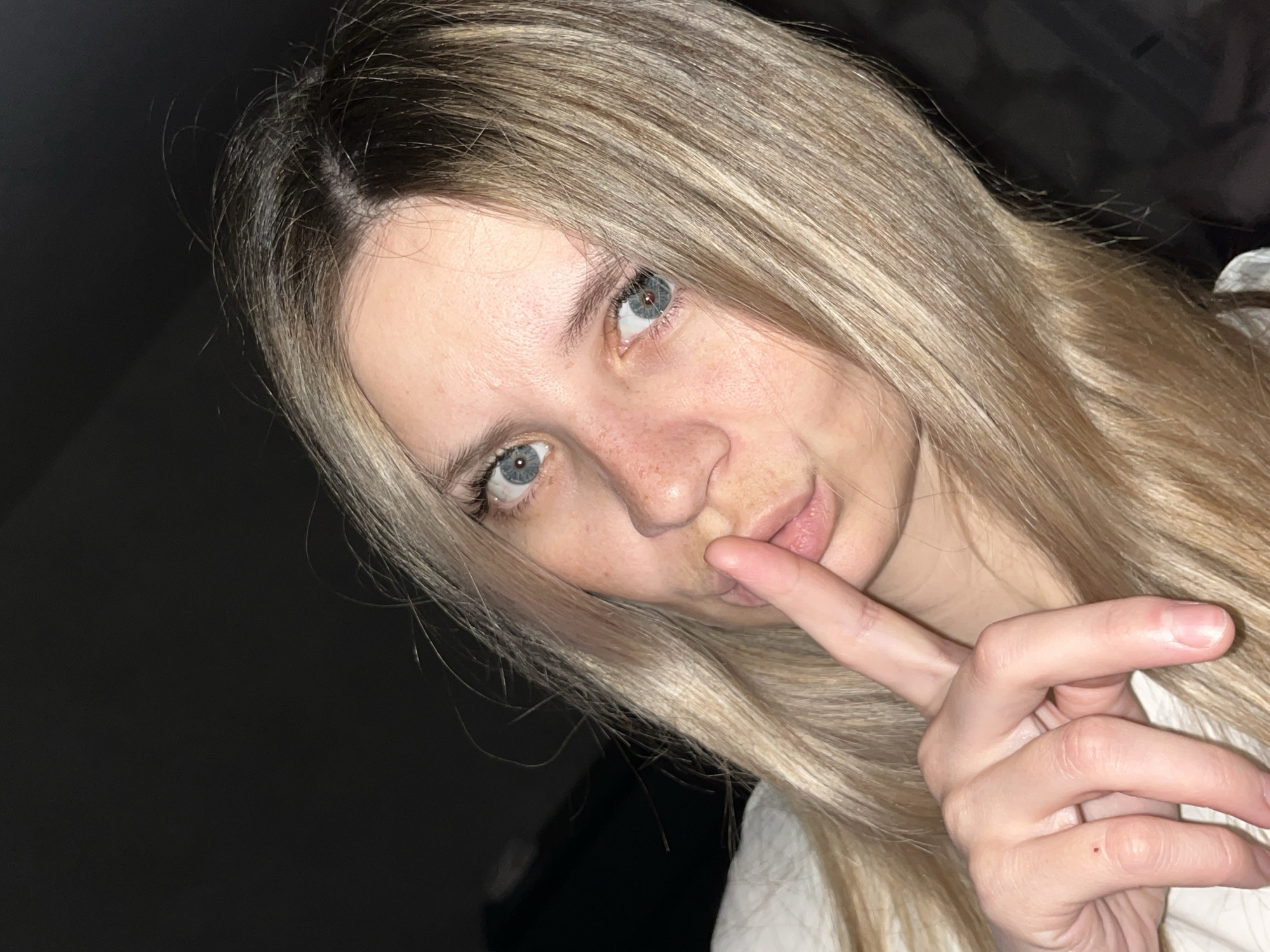A young woman with long, blonde hair and blue eyes holds a finger to her lips. She is fair-skinned with visible freckles. A hand and a white fabric or garment are visible in the foreground. The background is dark and indistinct. It is not possible to determine a specific location in Chaikovsky, Russia, from the given image.