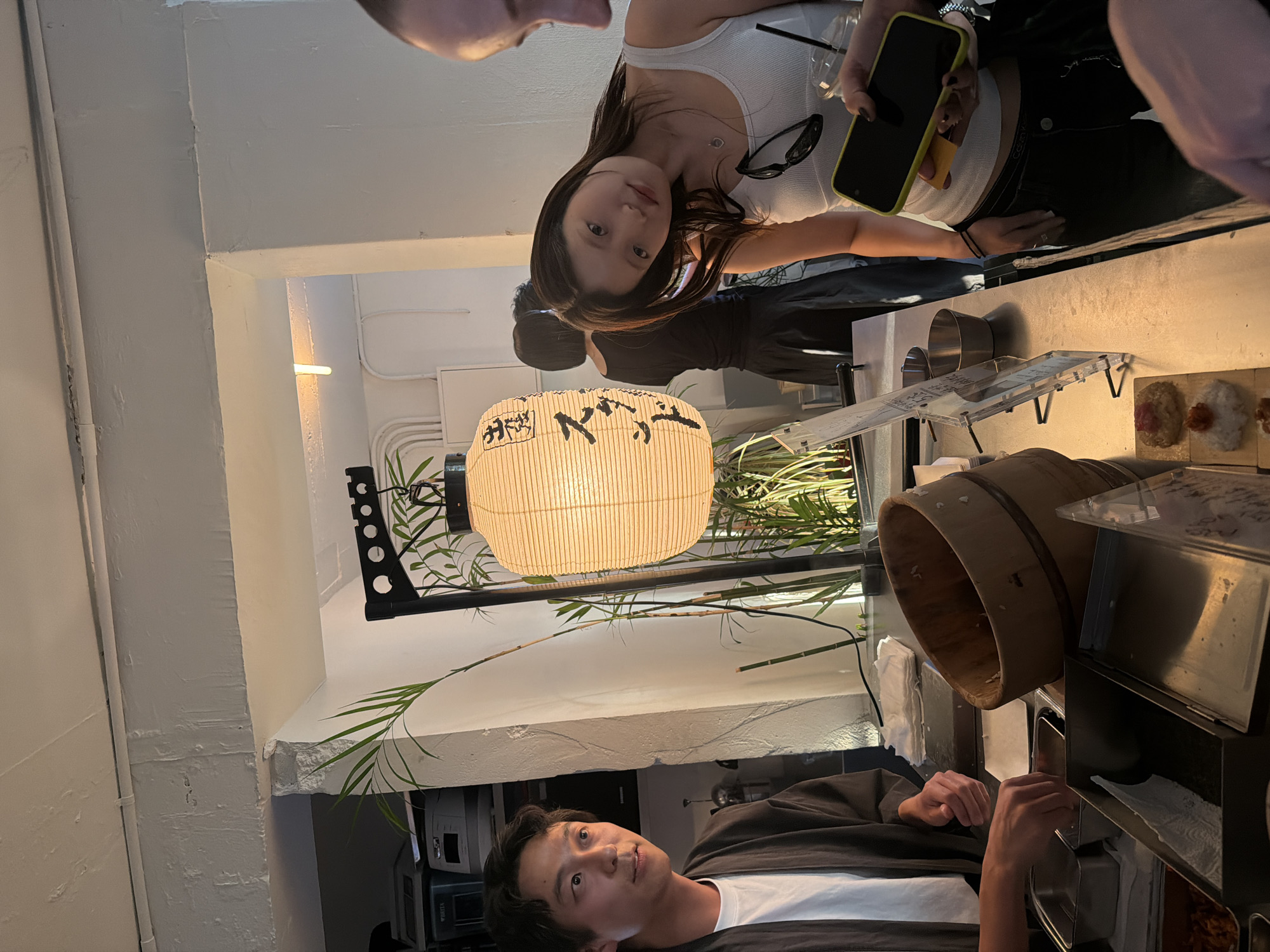 A medium shot captures a scene inside a restaurant, possibly located in Seoul, South Korea. An Asian man, dressed in a black jacket over a white t-shirt, is positioned at the lower portion of the frame. His gaze is directed upwards towards a woman with dark hair who is holding a smartphone and a beverage. The woman is wearing a white tank top and black pants. The restaurant appears to be a sushi establishment, with a wooden bucket and a sushi display case visible. Above, a traditional paper lantern provides illumination, and potted plants contribute to the ambiance. A second Asian man is seen in the background.