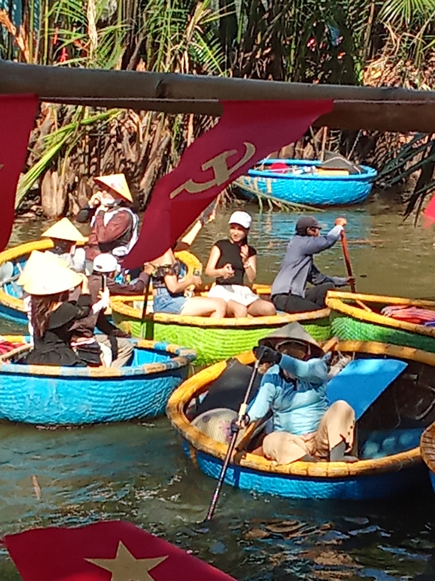 The image depicts a scenic canal in Da Nang, Vietnam, likely a tourist attraction, where people are enjoying rides in traditional Vietnamese basket boats. The boats, a distinctive round shape, are painted in vibrant blues and greens, with yellow accents along their rims. Red flags with a golden star, indicative of Vietnamese national symbols, are hung above the water, adding a festive touch.
Several individuals are visible within the boats. In the foreground, a person wearing a conical hat and a blue shirt is paddling a boat. To their right, another boat carries a person in a blue shirt and tan pants, also paddling. In the middle ground, a woman in a black top and white skirt is seated in a boat, smiling. Behind her, another person in a conical hat and brown attire is visible. Several other people in conical hats are also present in the boats, some appearing to be tourists. The water is calm, with reflections of the surroundings. Lush green vegetation lines the banks of the canal, suggesting a natural, tropical environment. The overall impression is one of a lively and picturesque tourist activity in a Vietnamese waterway.